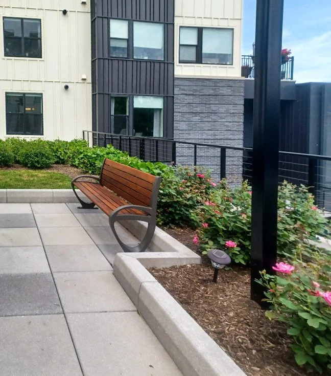 Urban outdoor seating area with a wooden bench on a tiled patio, surrounded by pink flowers and green bushes, near modern apartment buildings.