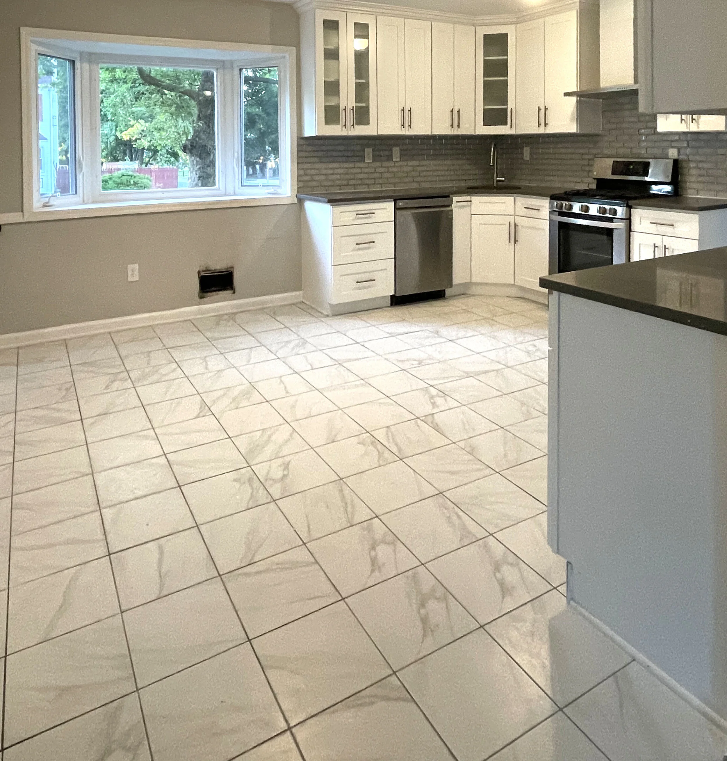 Clean kitchen with white cabinets, black countertops, stainless steel appliances, tiled flooring, and large window providing natural light.