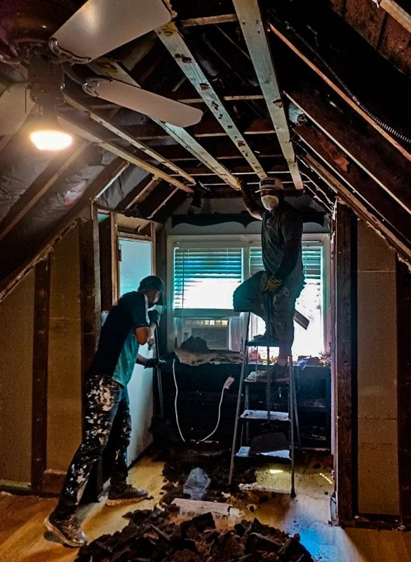Two people working on a room renovation, with exposed ceiling beams and insulation. One person is standing on a ladder while the other is using a tool near the window. Debris is on the floor.