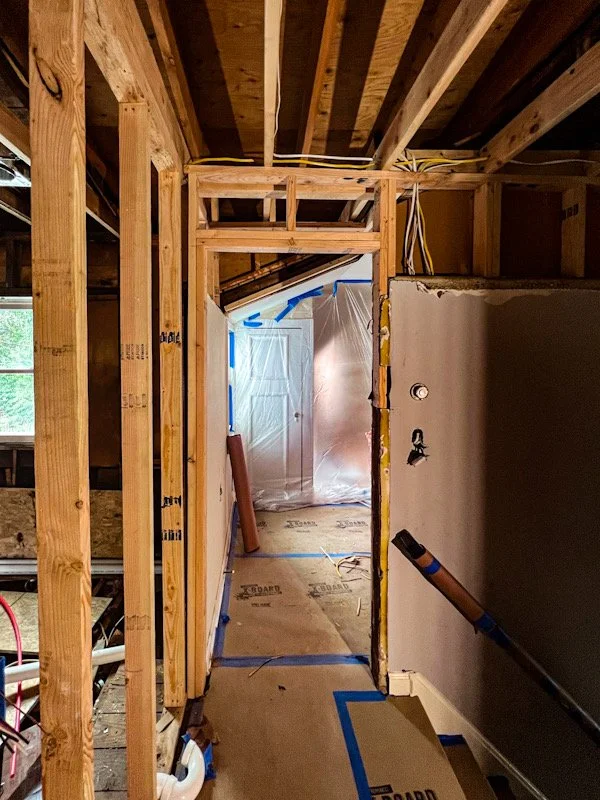 Interior view of a house under construction or renovation, featuring exposed wooden framing, electrical wiring, and partially finished walls with blue painter's tape and protective plastic covering on the adjacent room.