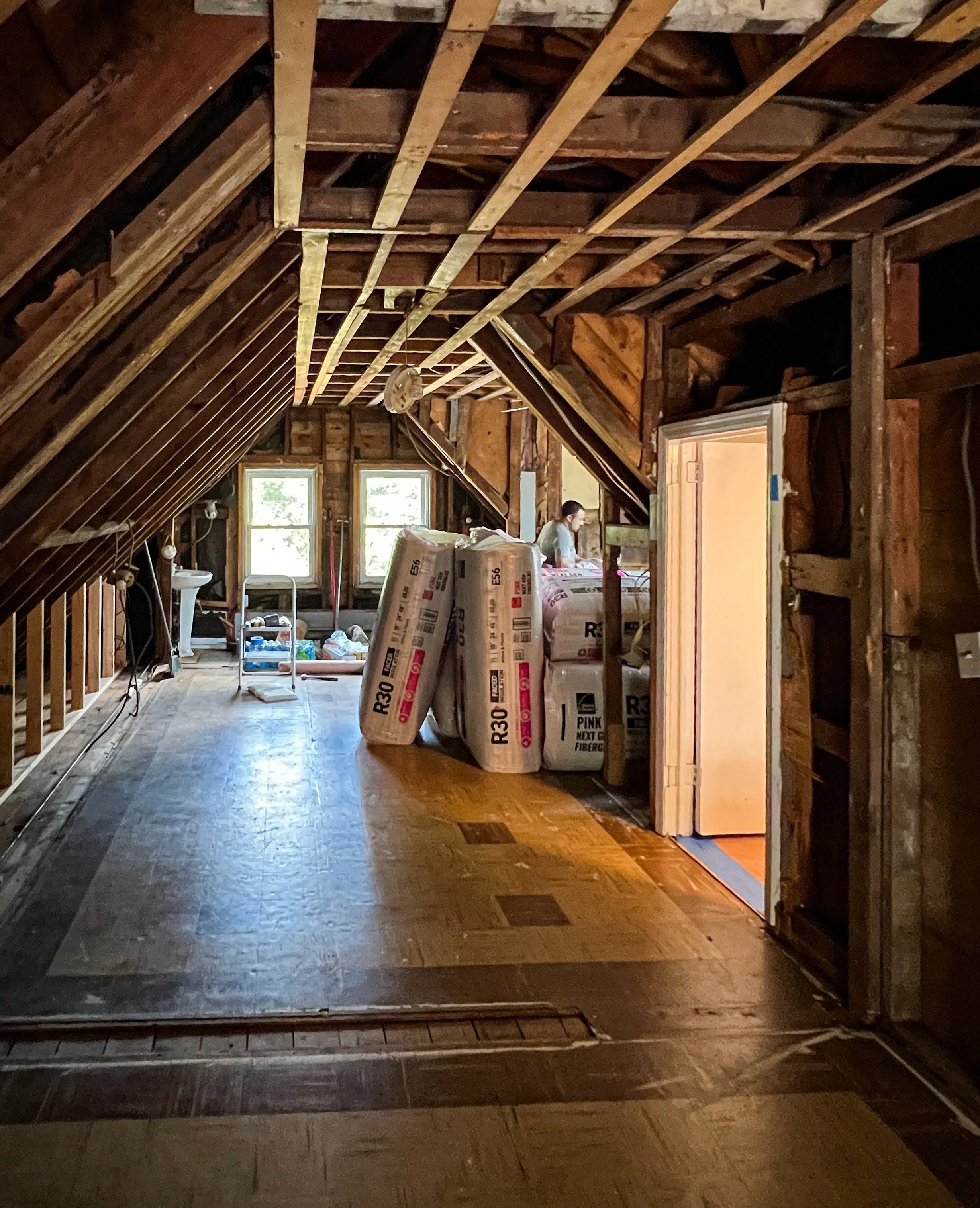Attic space under renovation with exposed wooden beams, bags of insulation, and a woman working near the windows.