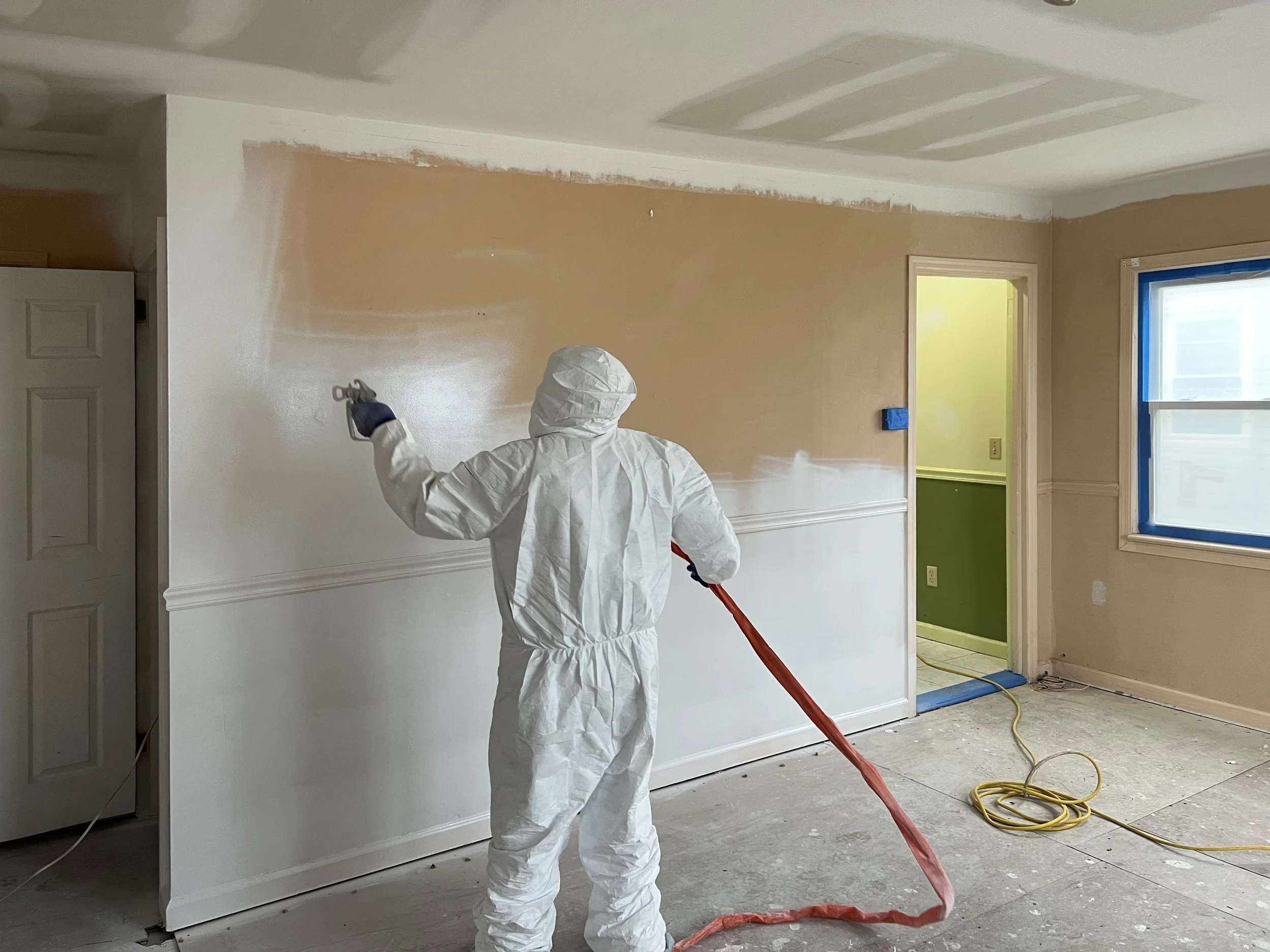Person in white protective suit spray painting a wall in a room under renovation.