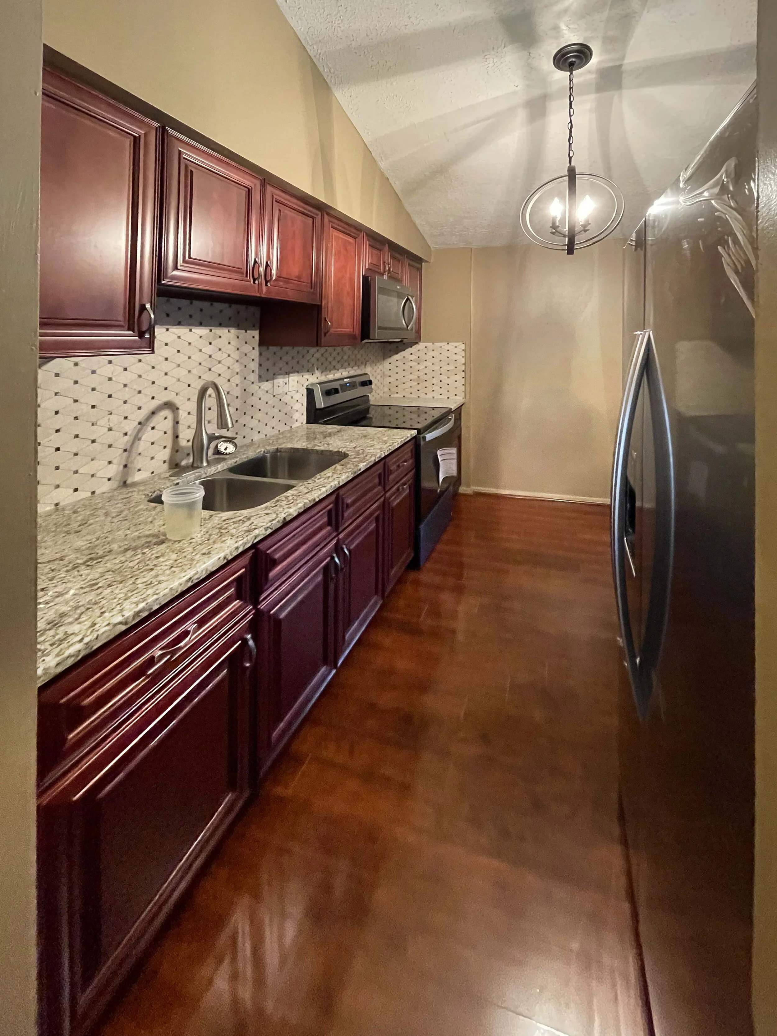 Kitchen with dark wood cabinets, granite countertop, mosaic tile backsplash, stainless steel sink, black stove, microwave, and refrigerator, with wood flooring and a hanging light fixture.