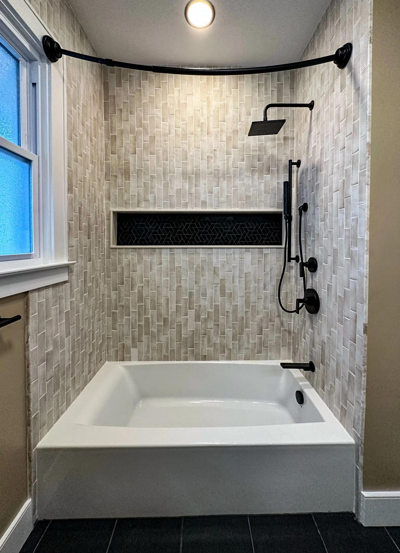 Modern bathroom shower with beige tile walls, a built-in black tile niche, black shower fixtures, a window, and a white bathtub.