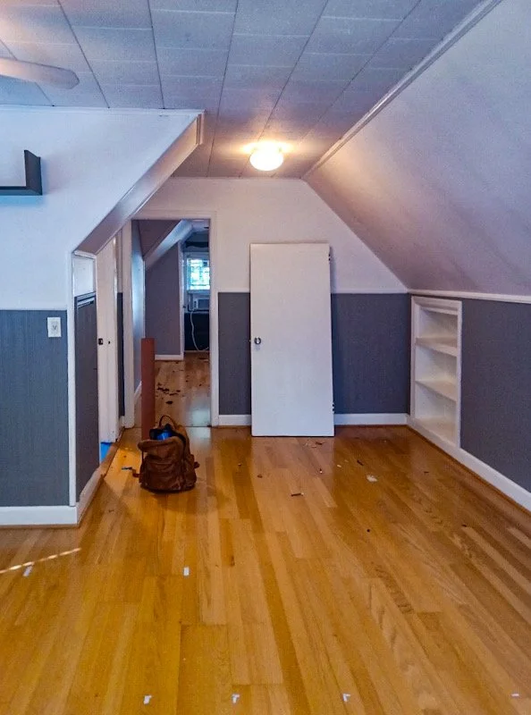 Empty room with hardwood floors, sloped ceiling, white walls with gray wainscoting, a small white door, built-in shelves, a backpack on the floor, and a ceiling light turned on.