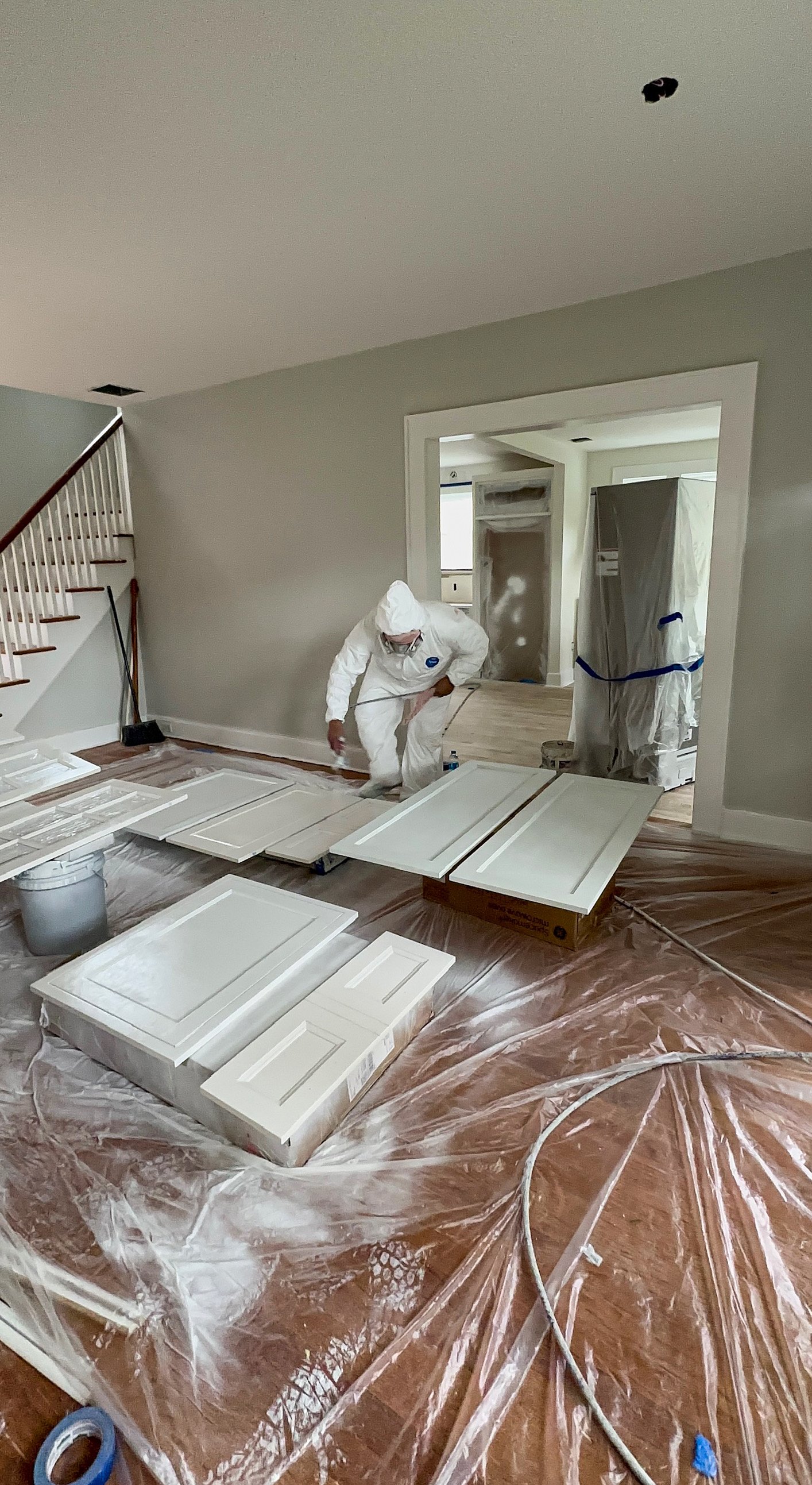 A person in white protective clothing is painting cabinet doors inside a house under renovation. The room has plastic covering on the floor and new cabinetry in the background.