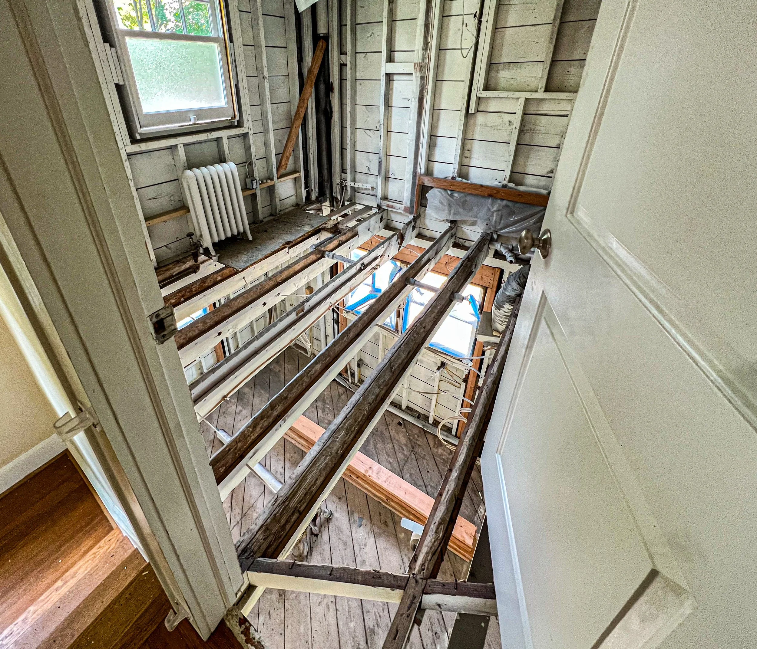 Interior view of a house under renovation, showing exposed wooden floor joists and framing in a room with a window, radiator, and construction materials.