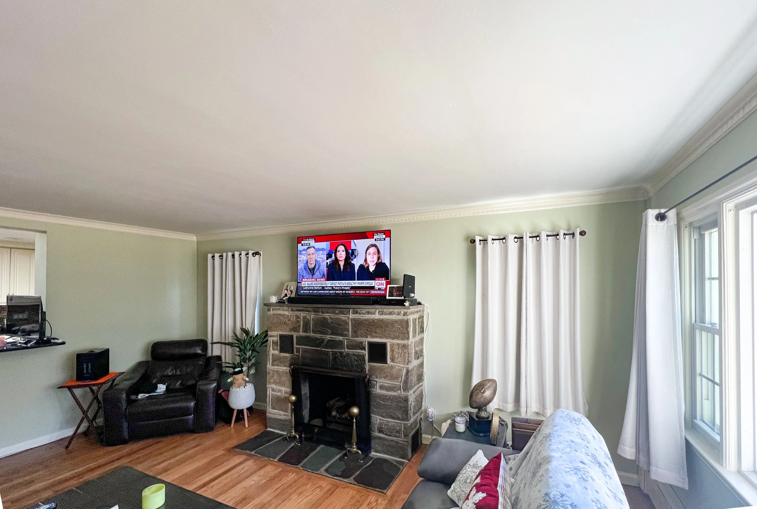 A living room with a stone fireplace, a mounted flat-screen TV, beige curtains, a black armchair, a potted plant, a white sofa, a small side table with sports equipment, and windows with white curtains.