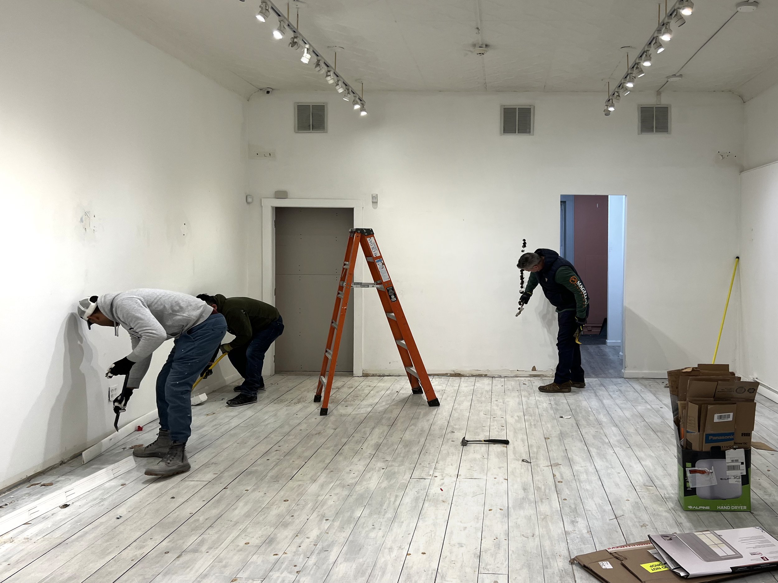 Three workers renovating a white-walled room, one using a level, one on the floor with a tool, and one with a drill, with construction materials and a ladder present.