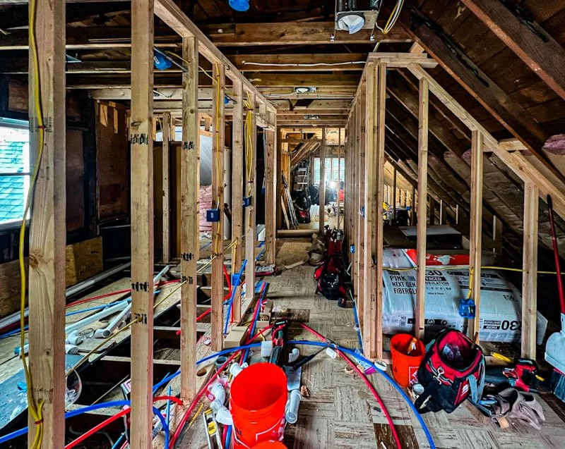 Interior view of a house under construction or renovation with exposed wooden framing, electrical wiring, and plumbing pipes.