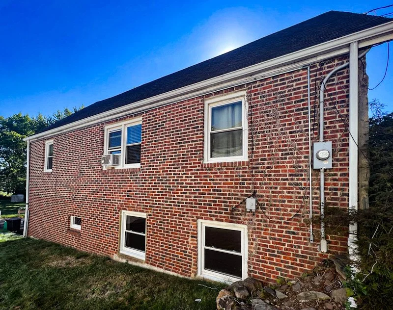 The side of a two-story brick house with five windows, including one with an air conditioning unit, and visible electrical meters and wiring.