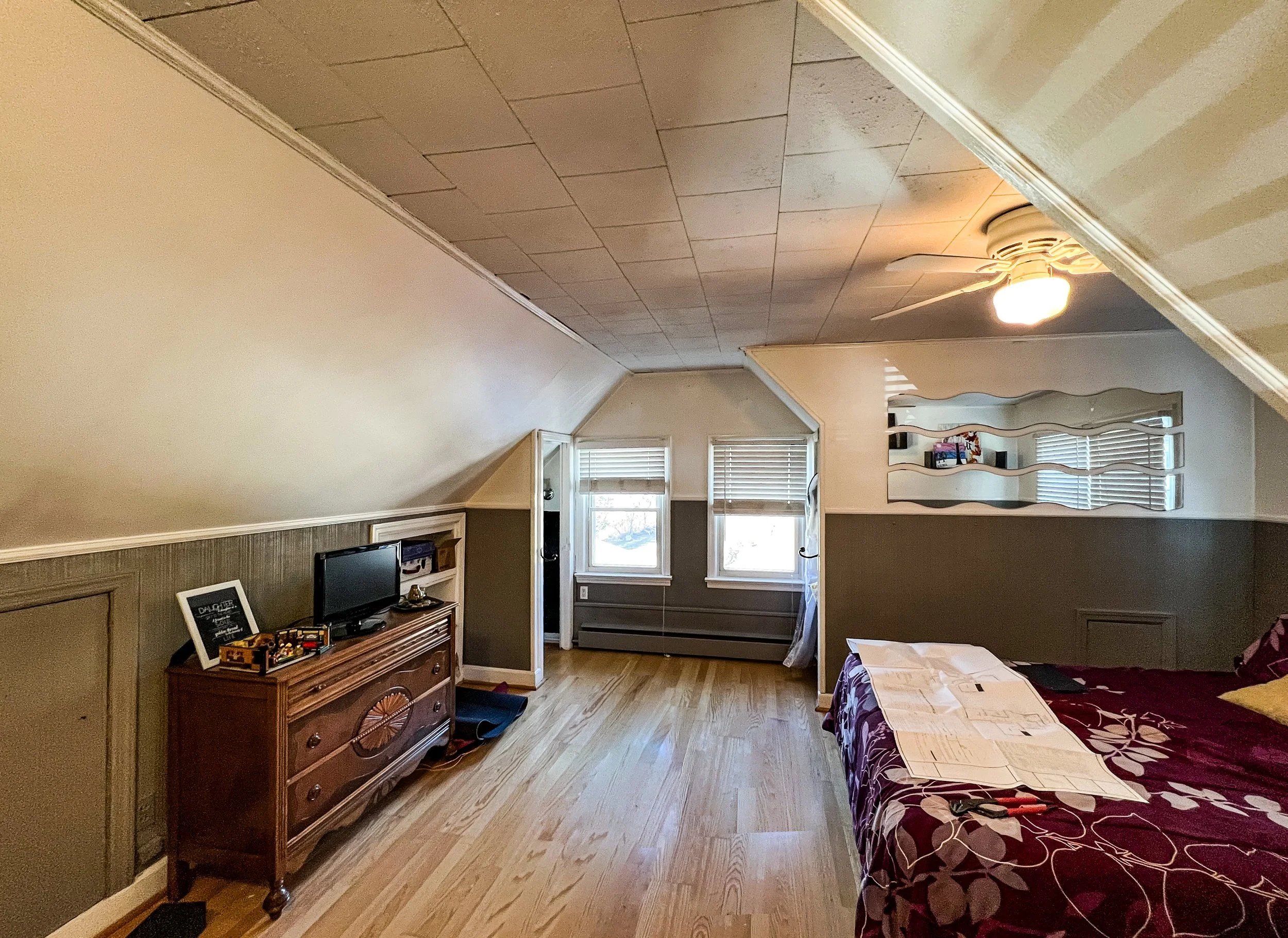 A bedroom with a sloped ceiling, a ceiling fan with light, a bed with dark purple bedding, a mirror on the wall, a small wooden dresser with a TV and decorative items, and two windows with blinds.