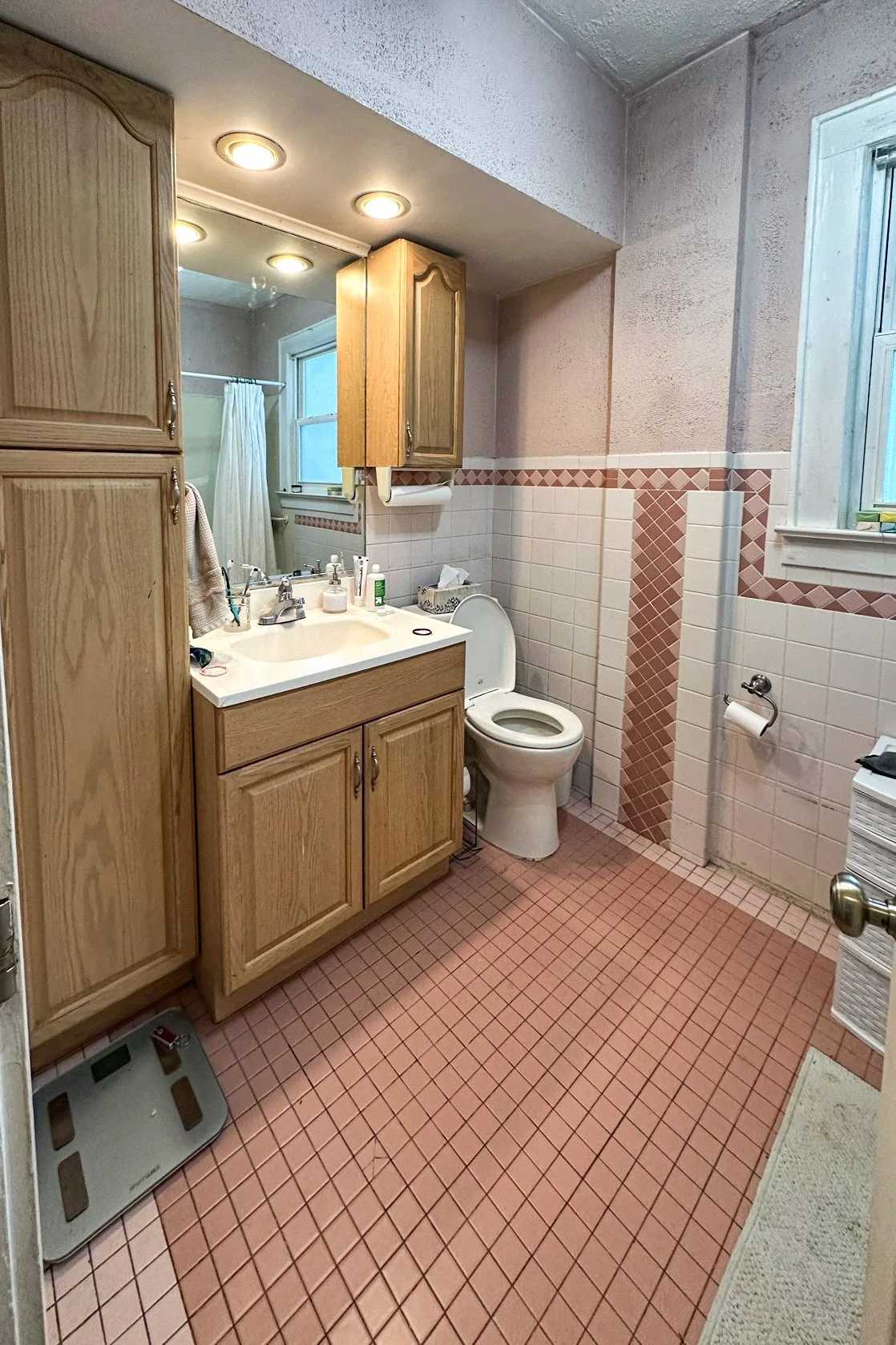 A small bathroom with pink tiled floor and half pink and white tile walls. A wooden cabinet with drawers and a mirror above the sink, and a small window with a white curtain. A toilet with a lid open, a toilet paper holder, and a window with frosted 