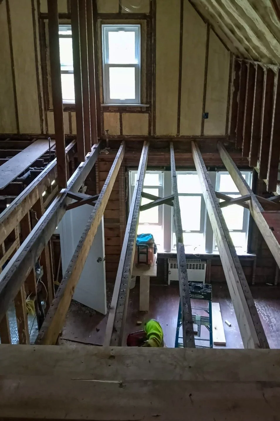 Interior of a house under renovation with wooden framing and two windows, showing exposed beams and construction tools.