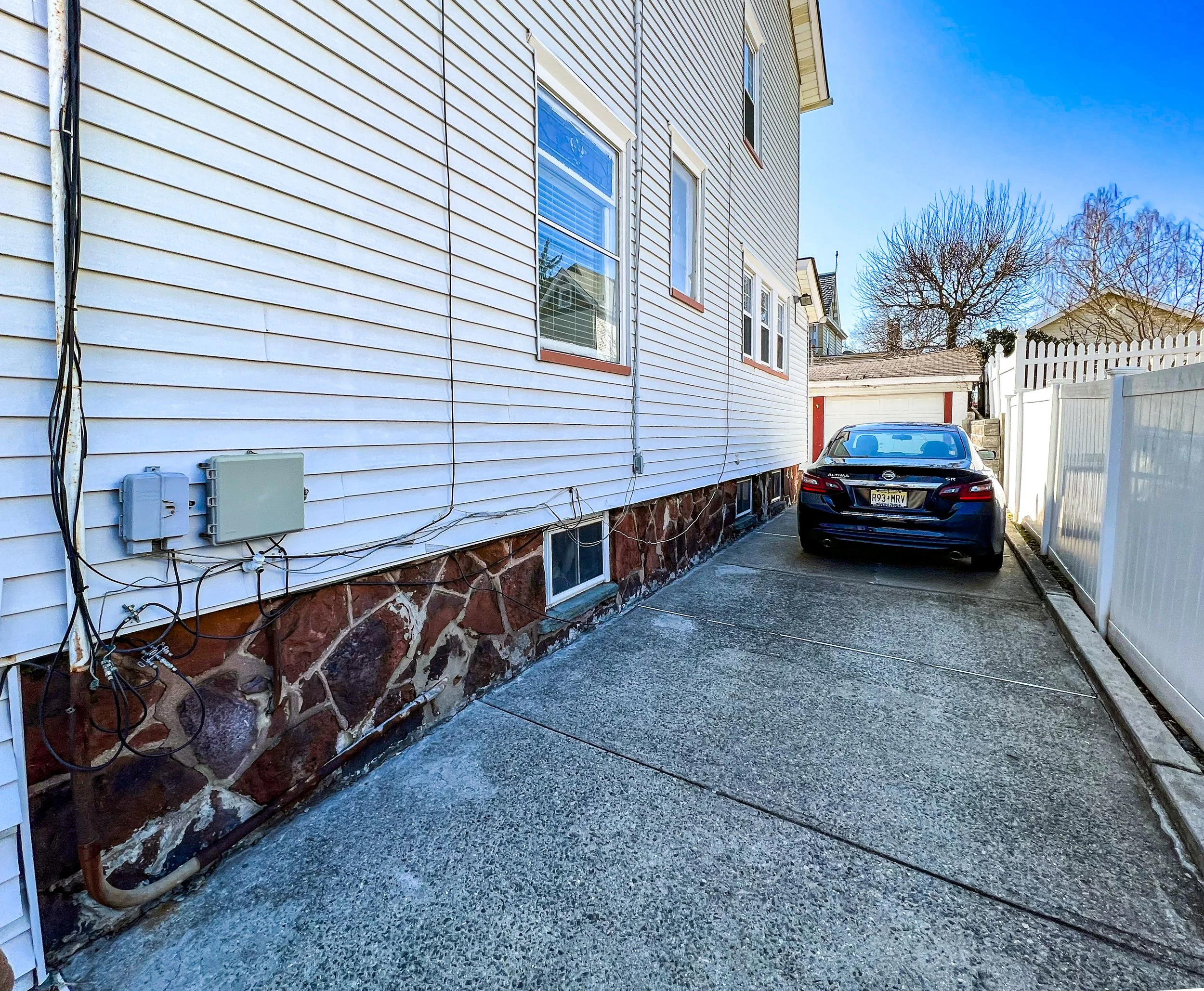 A concrete driveway next to a white house with basement windows, with a black car parked near a white fence. The house has vinyl siding and a stone foundation, with visible utility meters and wires. The background includes trees, a garage, and a sunn