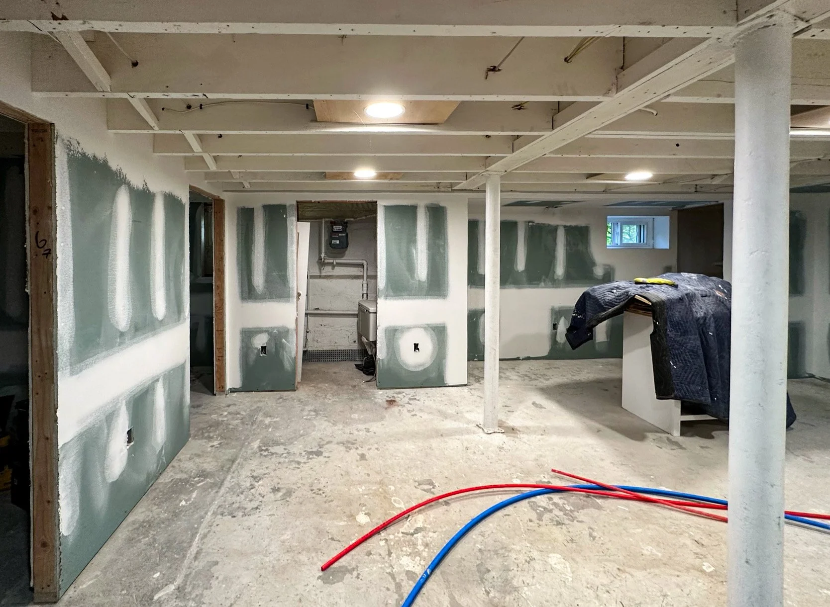 Interior of a basement under construction with drywall being installed, exposed ceiling beams, insulation, and construction materials on the floor.