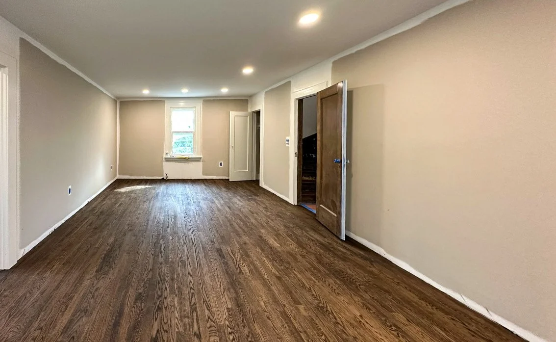 Empty living room with hardwood floors, beige walls, a single window, and doorways leading to other rooms.