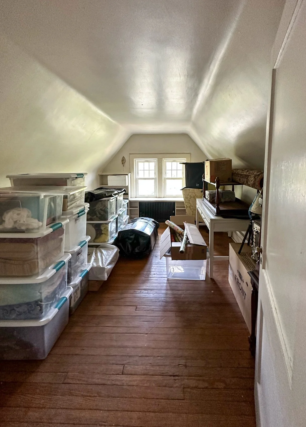 Cluttered attic with storage bins, boxes, and various household items, natural light from window at the end of the room.