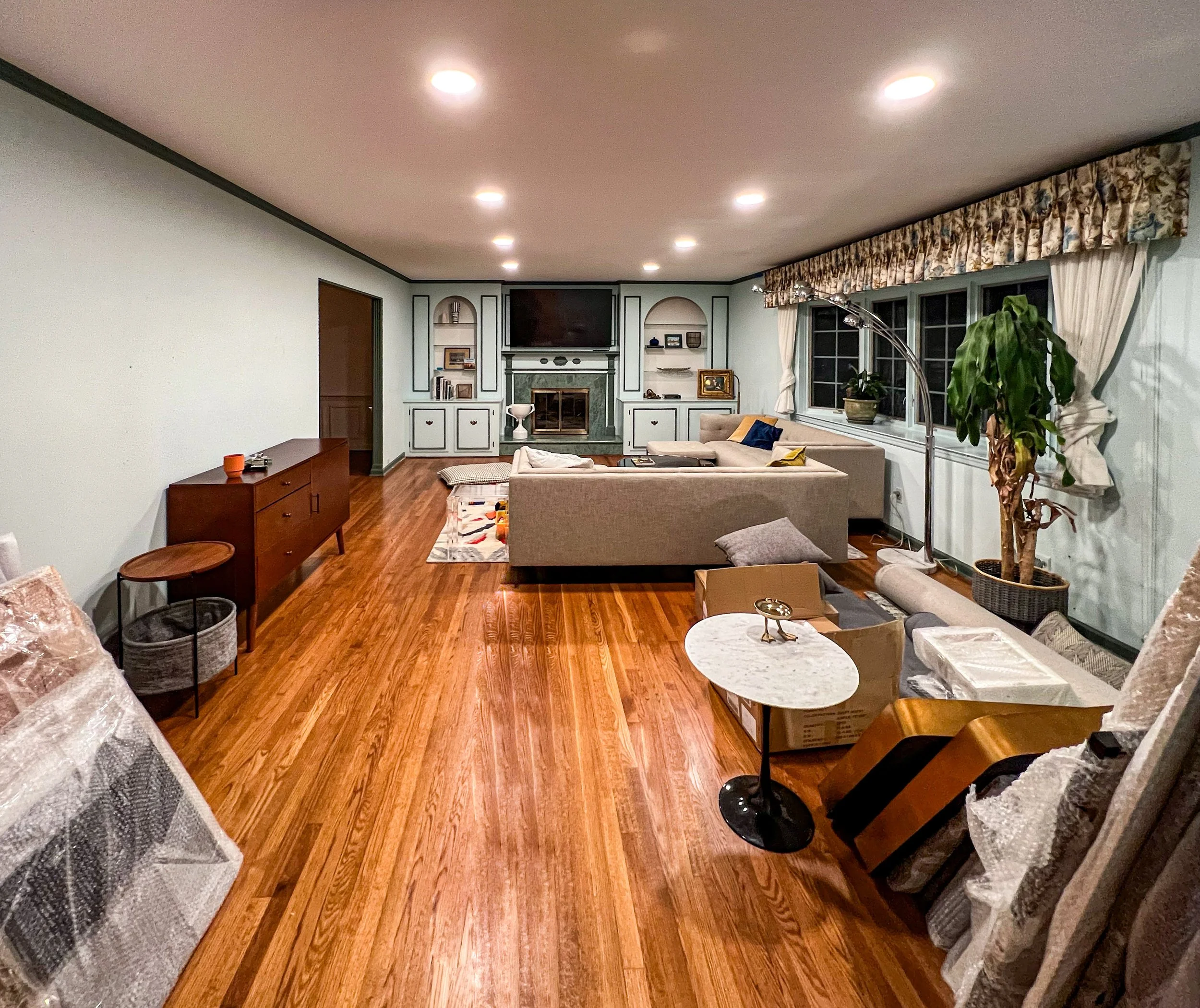 Living room with hardwood floor, beige sectional sofa, large window with floral valance, potted plant, TV above fireplace, and various pieces of furniture, some wrapped in plastic.