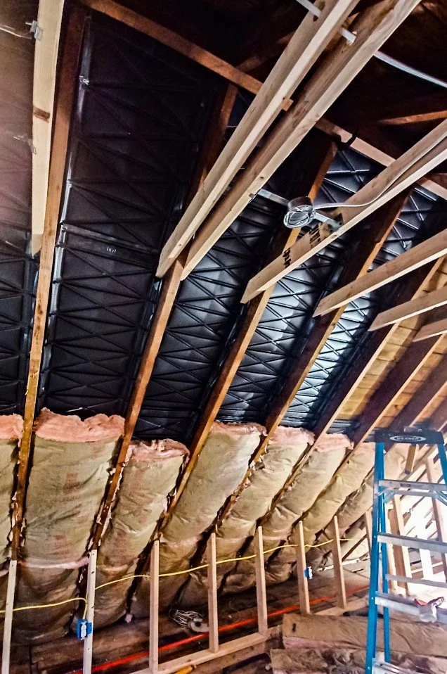 Insulation and framing being installed in an attic roof space, with angled rafters, black insulation boards, and fiberglass batt insulation along the lower part, and a blue step ladder on the right.