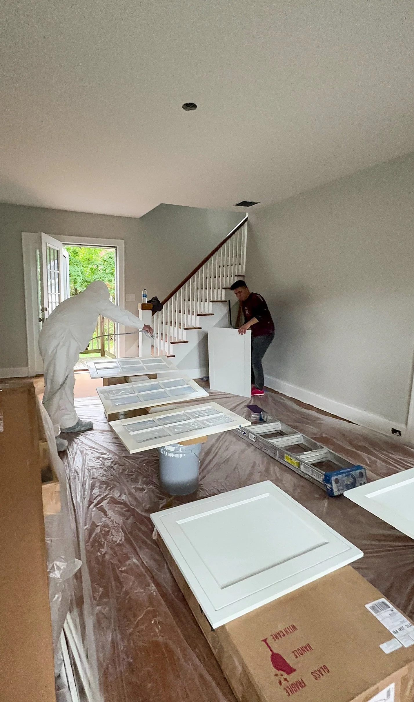Two workers are installing cabinets inside a house. One worker is in a protective suit, and both are handling cabinet doors. The room has a staircase, a door leading outside, and plastic sheeting on the floor.