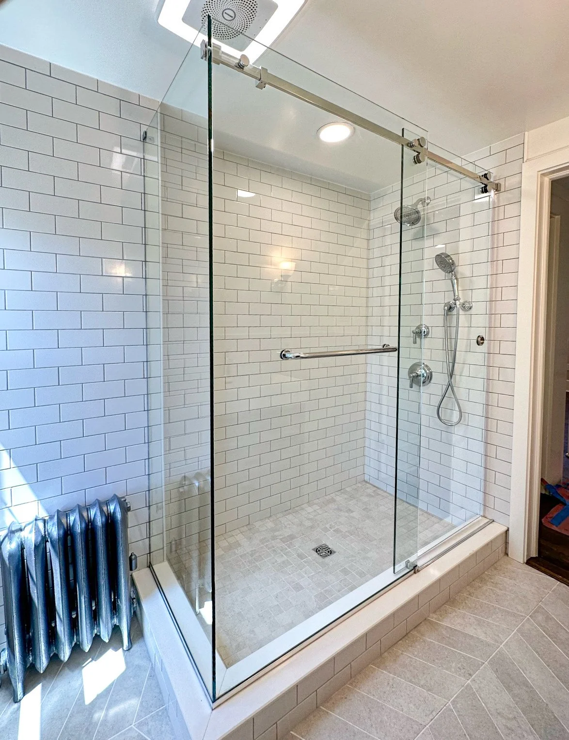 A modern walk-in shower with a glass enclosure, white subway tile walls, a rain showerhead, a handheld showerhead, and a small tiled shower floor.