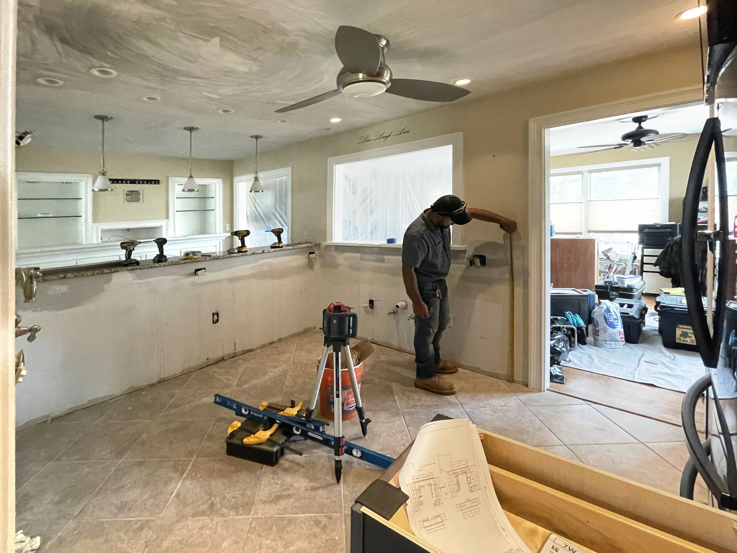 A man wearing a gray shirt and headphones measures the wall in a kitchen under renovation. The room is partially finished with open electrical outlets, tools on the counter, and construction plans on a table. Adjacent rooms show tools and supplies.