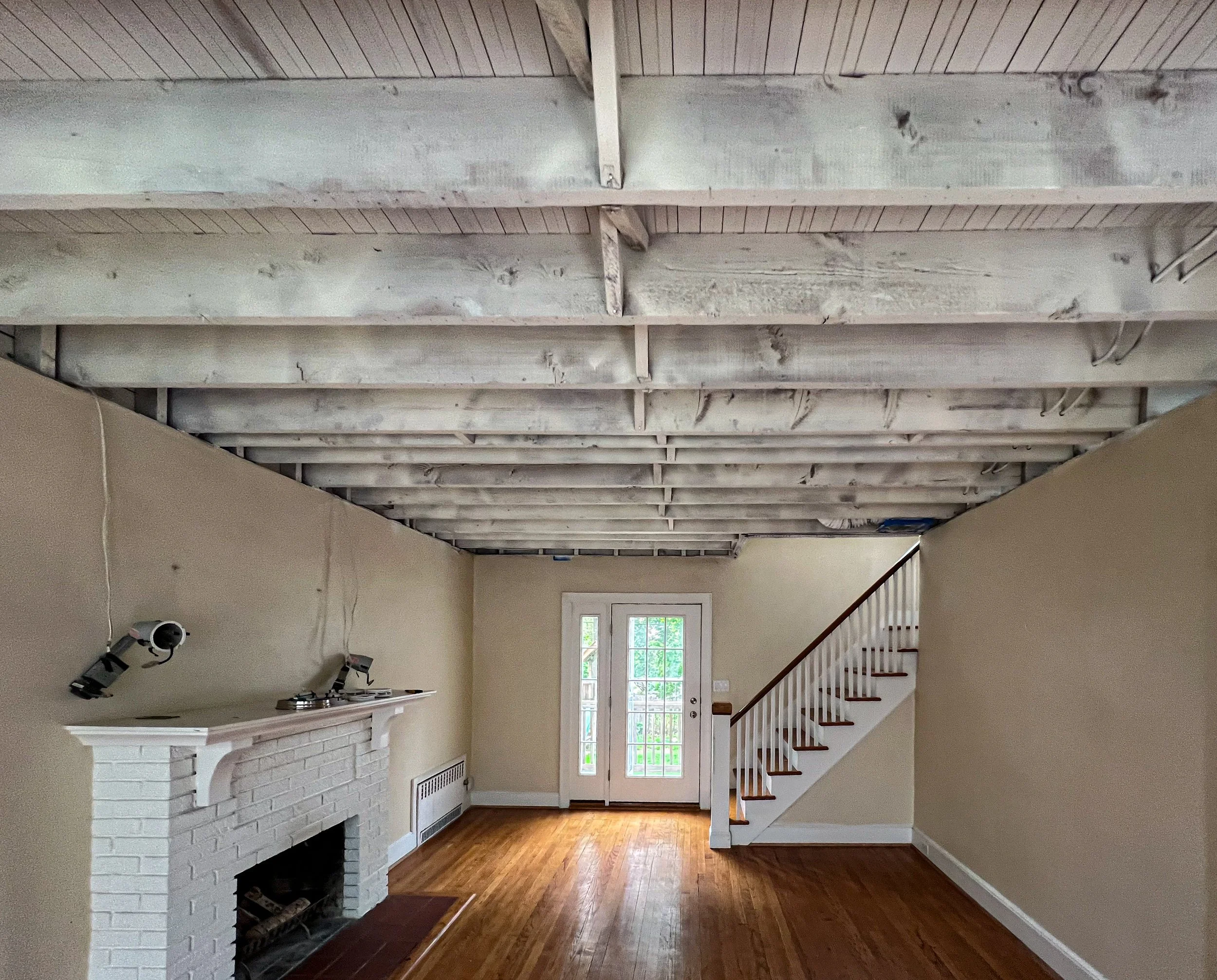 Living room with an unfinished ceiling where the beams are exposed, a white brick fireplace, hardwood floors, a set of glass-paneled double doors, a staircase with white railing on the right, and beige walls.