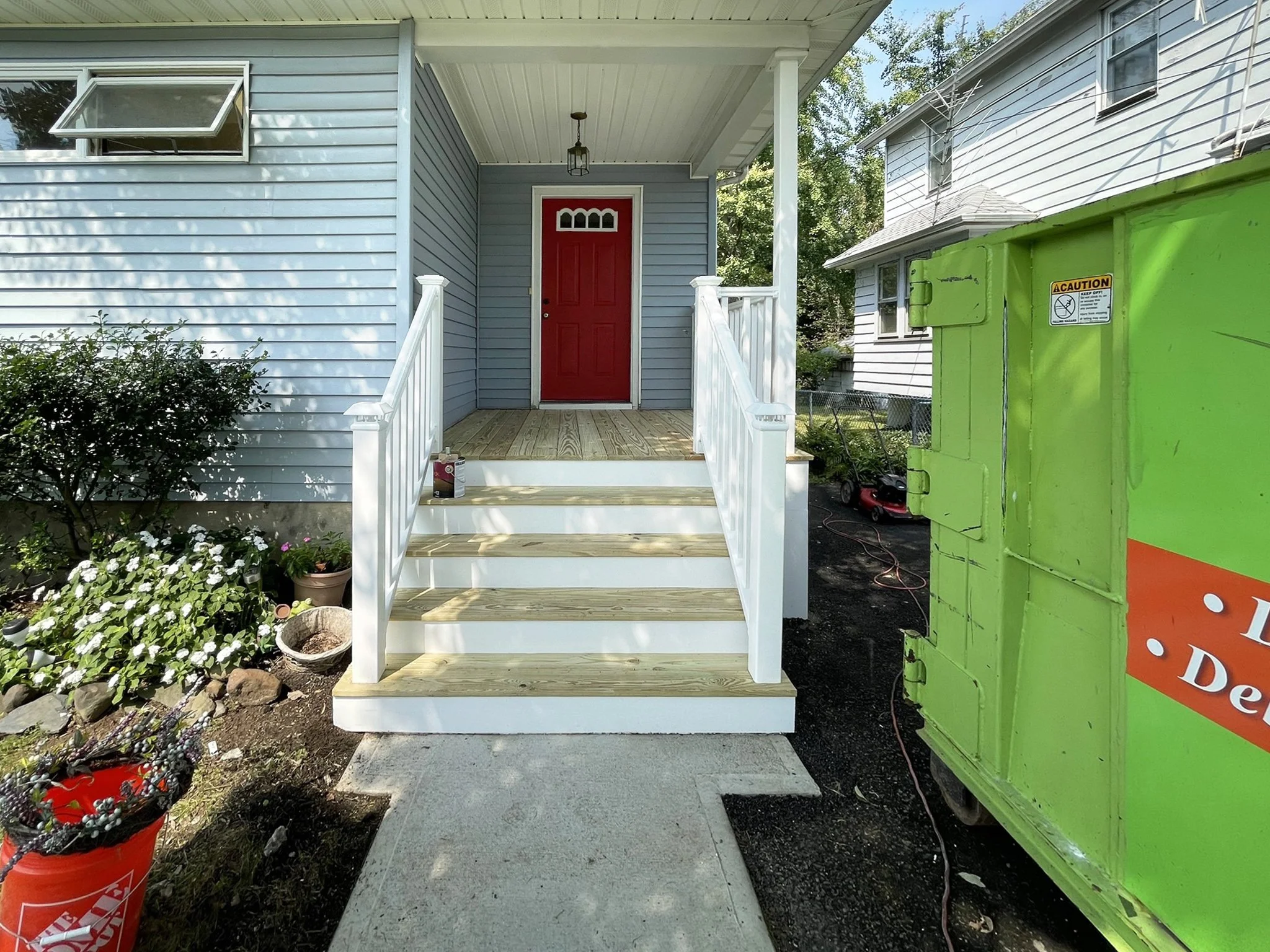 Front porch of a house with a bright red door, white stairs and trim, light blue siding, a small porch ceiling light, and garden plants on the left side. There is a green utility box on the right side, and a concrete walkway leading up to the stairs.