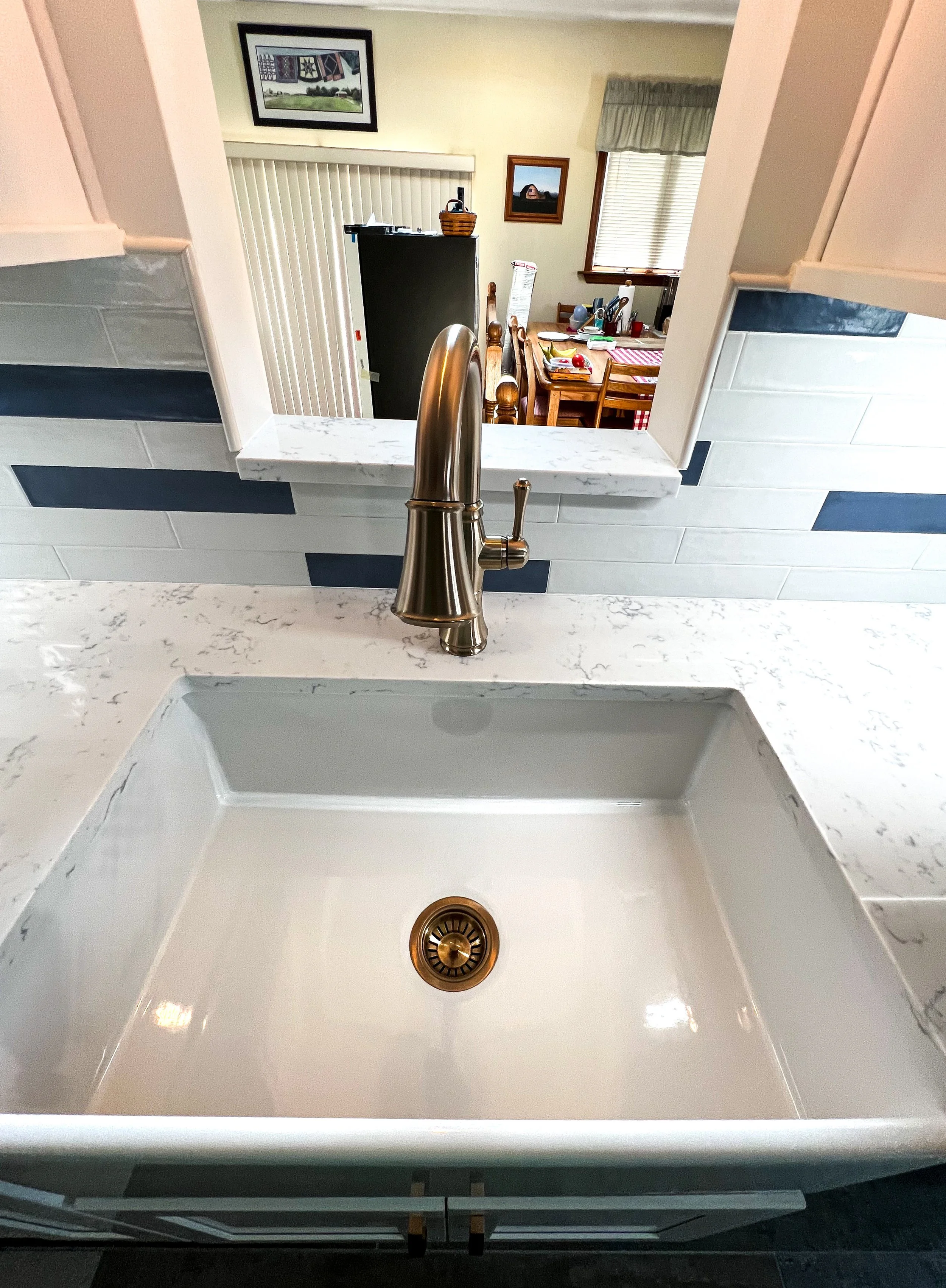 Kitchen sink with a brushed metal faucet, white marble countertop, and a view into the dining area with a wooden table and chairs through a pass-through window.