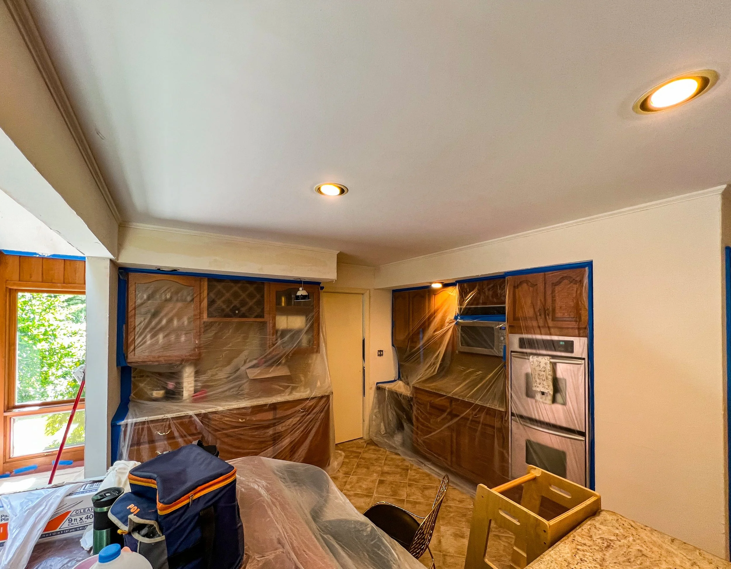 Kitchen undergoing renovation, with cabinets and appliances covered in plastic, a window on the left, and various tools and materials on the counter and table.