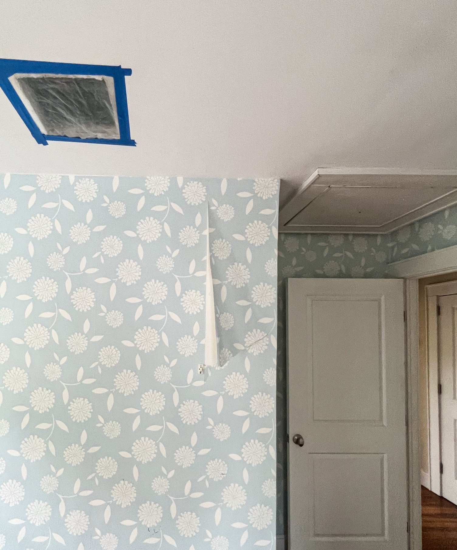 Part of a room undergoing renovation with a section of wallpaper on the wall, a door slightly open, and an unfinished ceiling vent with blue painter's tape around it.