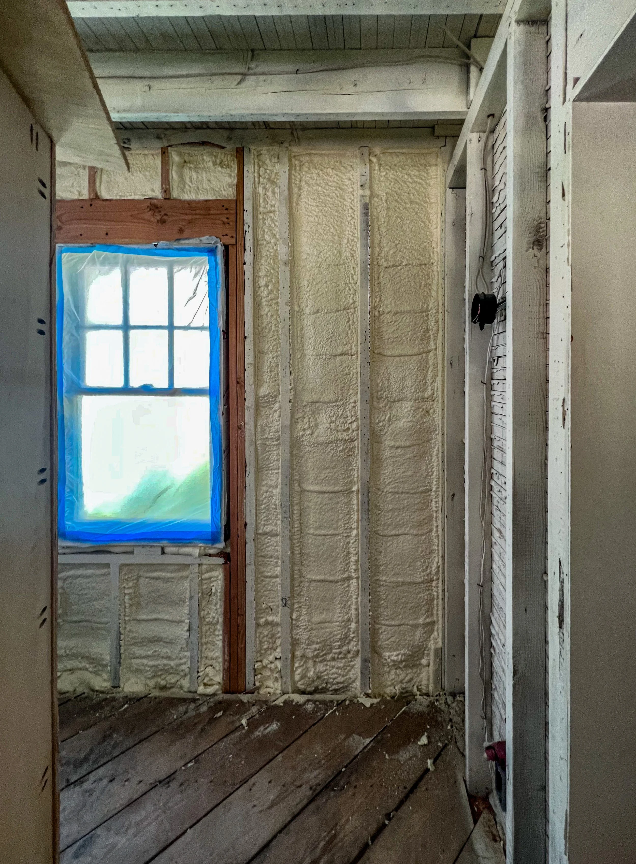 Interior of a room under renovation with a window covered in green plastic, spray foam insulation on the walls, and exposed wooden framing.