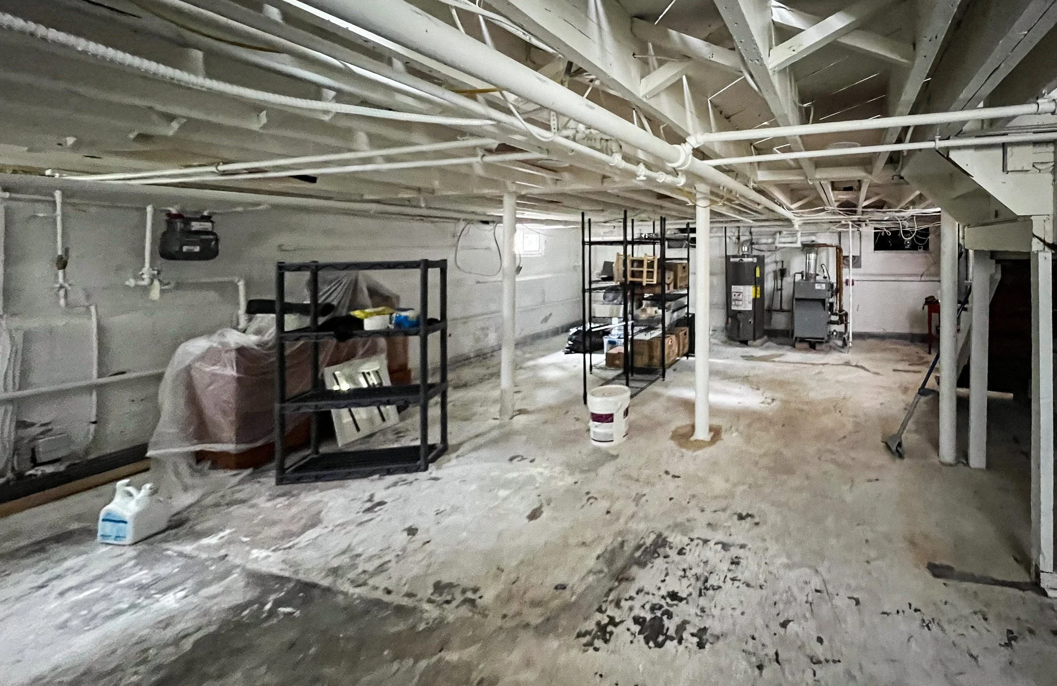 An unfinished basement with exposed pipes, concrete walls, and construction supplies, including shelving units, a bucket, and tools.