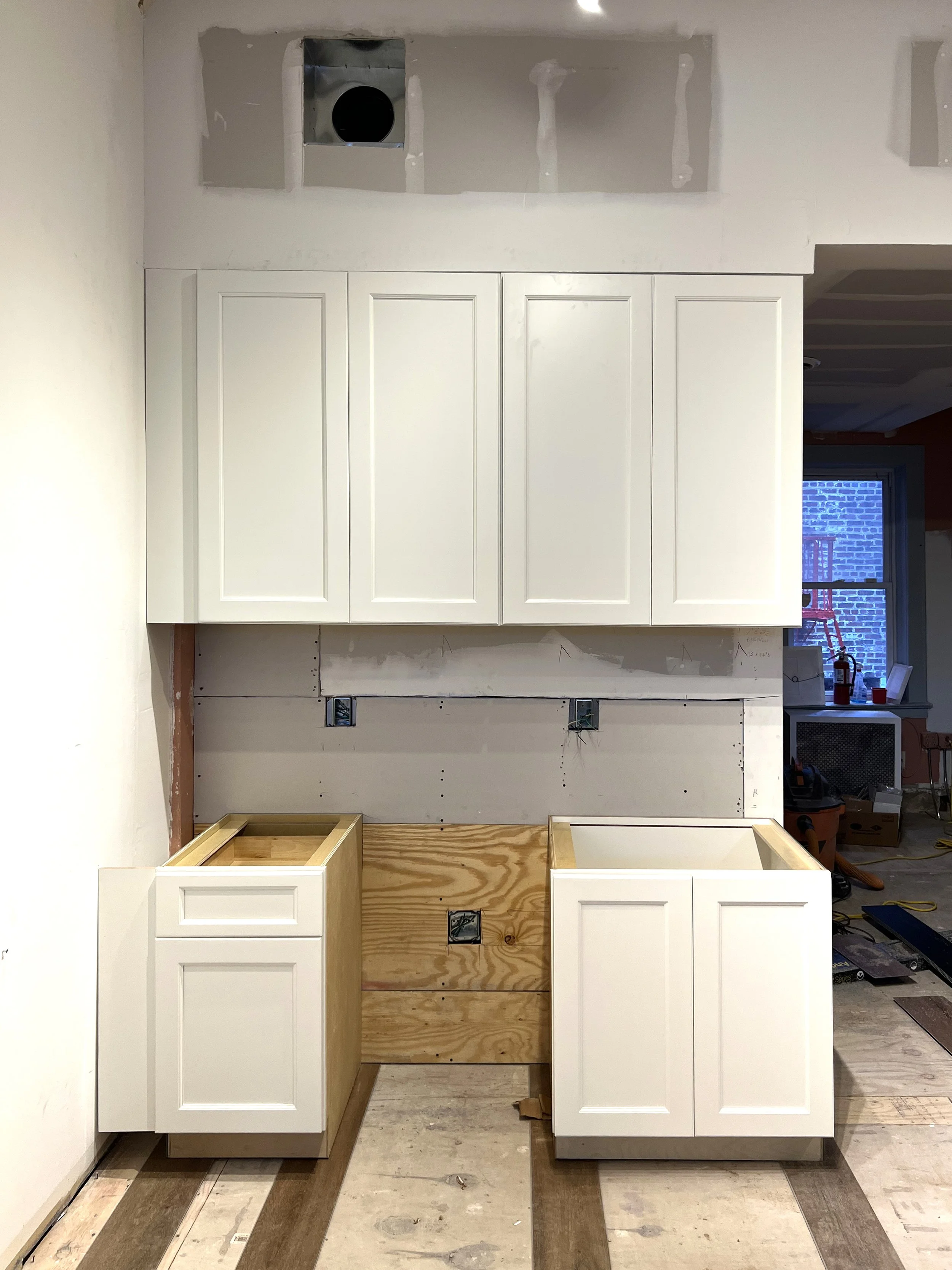 Kitchen cabinet installation in progress with upper white cabinets and lower wooden cabinets being set up, construction tools and materials visible.