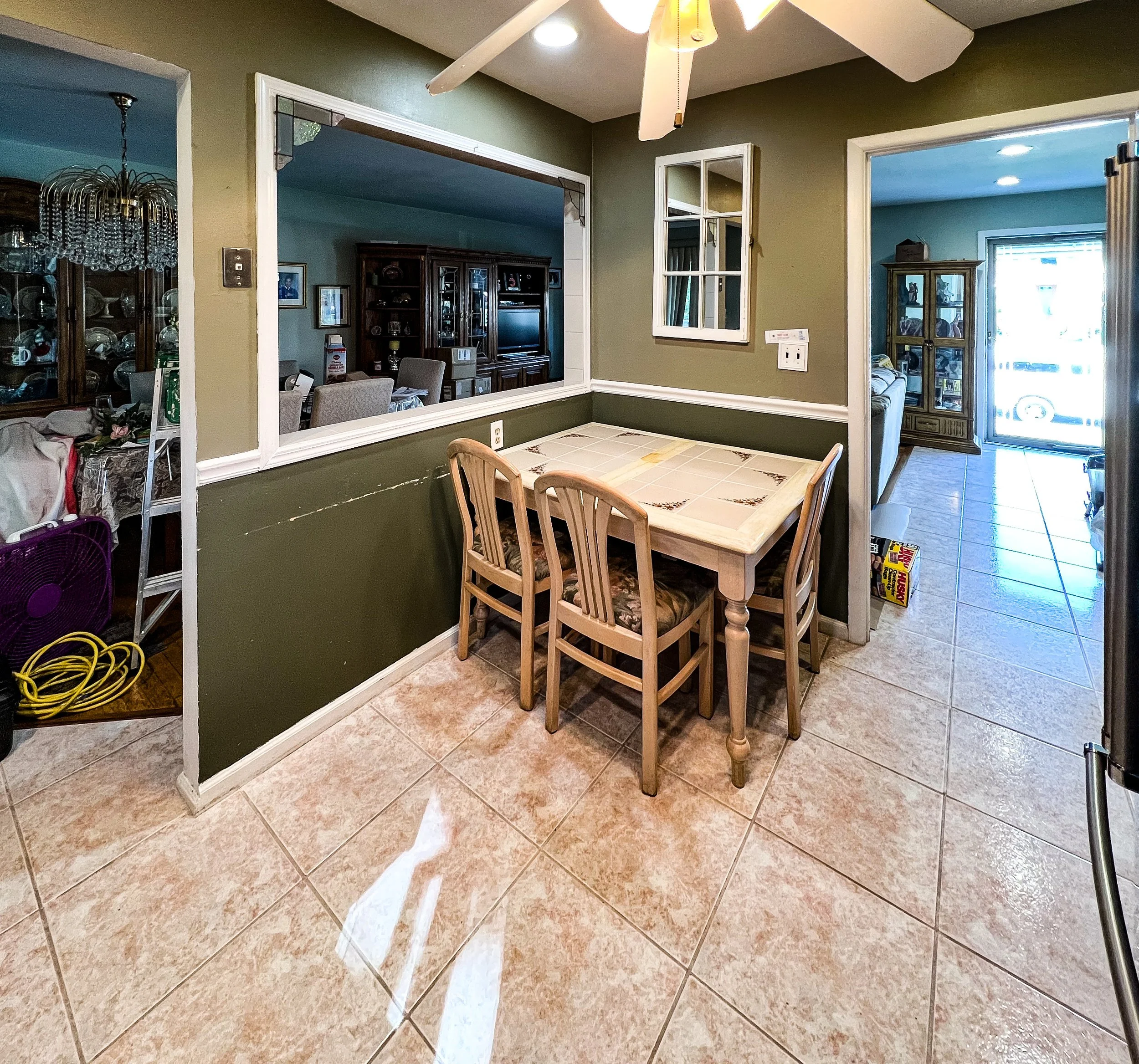A dining area with a wooden table and four matching chairs, separated from a living room by a half wall with a large opening, in a home with tiled floors and green walls.