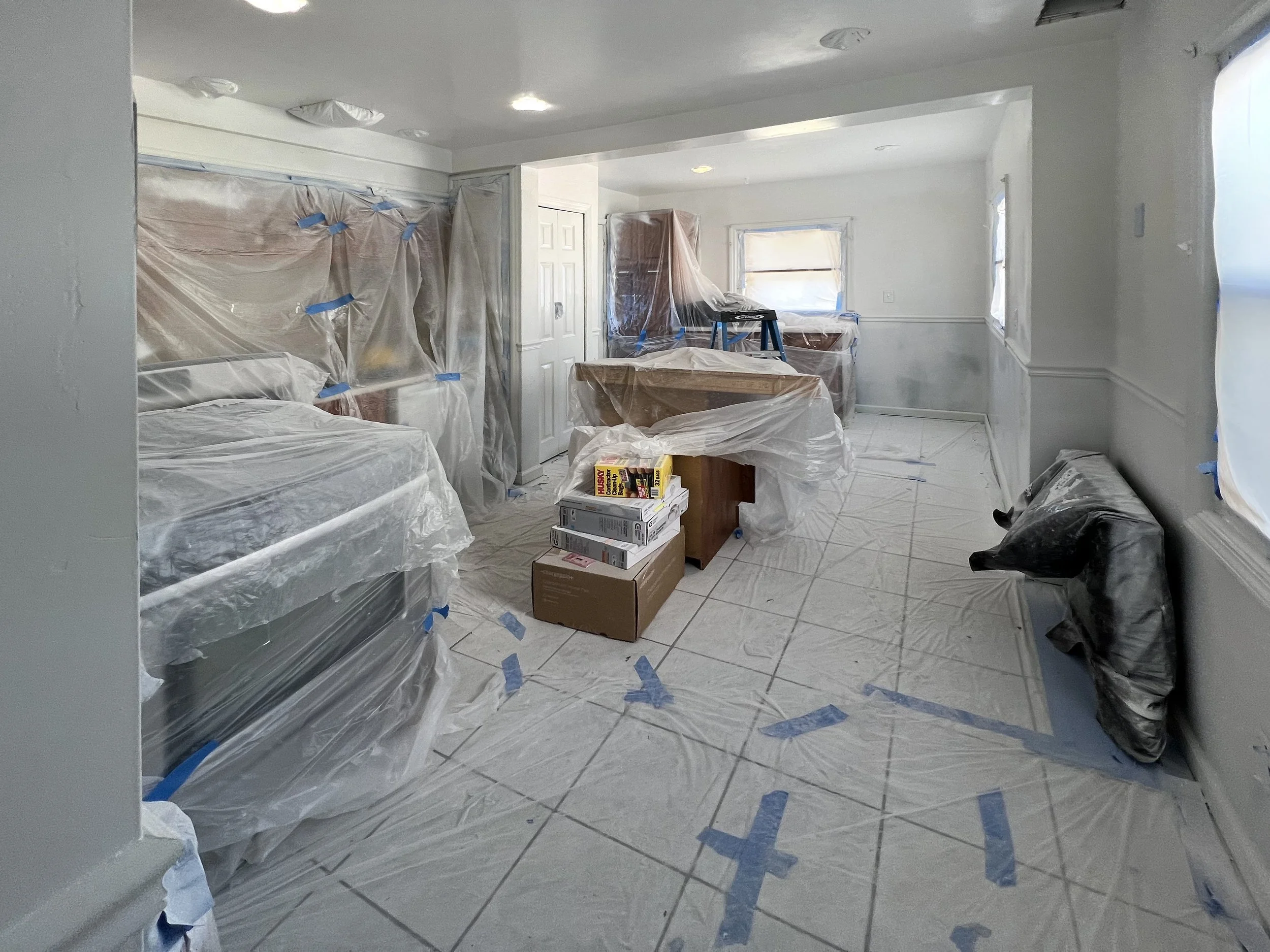 A room under renovation, with furniture covered in plastic and blue painter's tape, boxes and tools on the floor, and windows with plastic coverings.