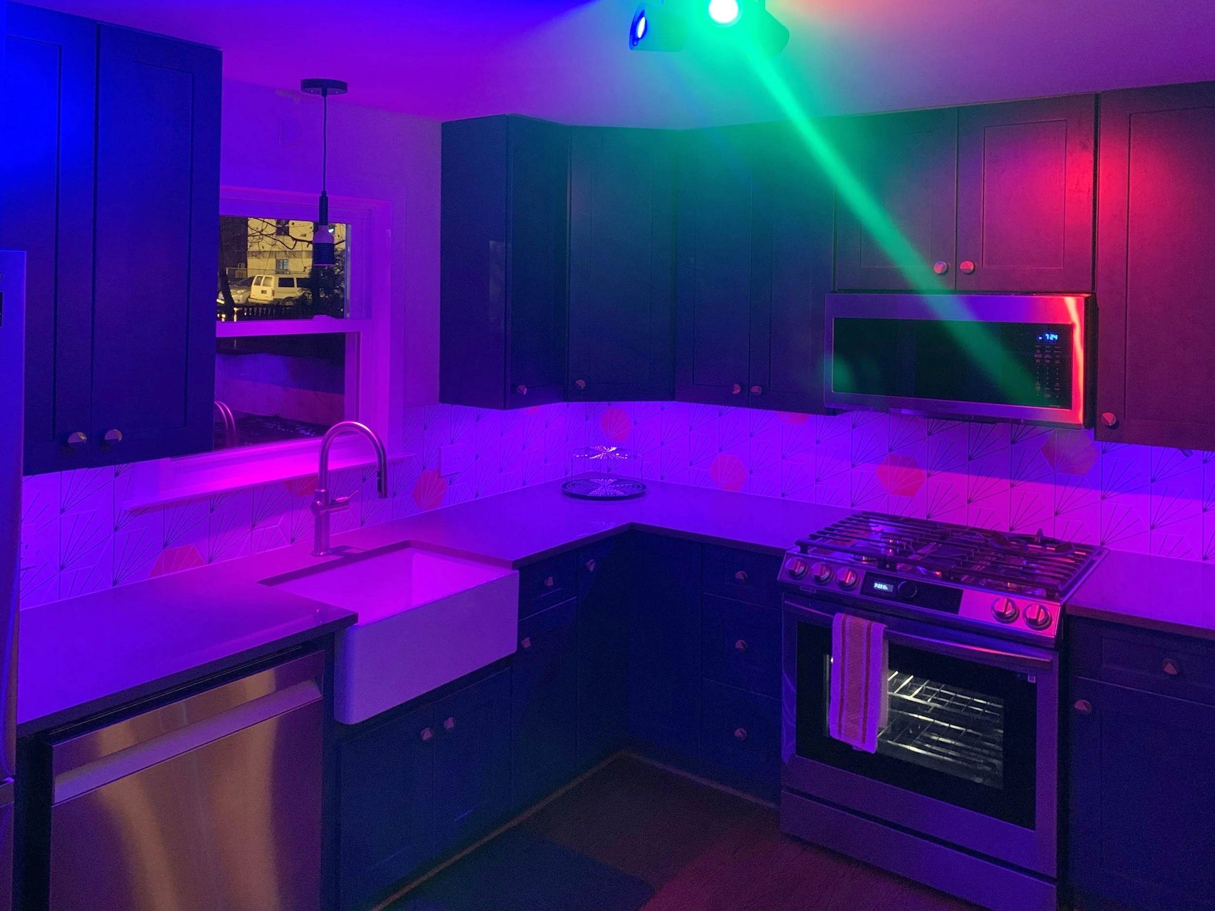 A kitchen illuminated with colorful laser lights, with dark blue cabinets, a white farmhouse sink, a stovetop, microwave, and retro patterned backsplash.