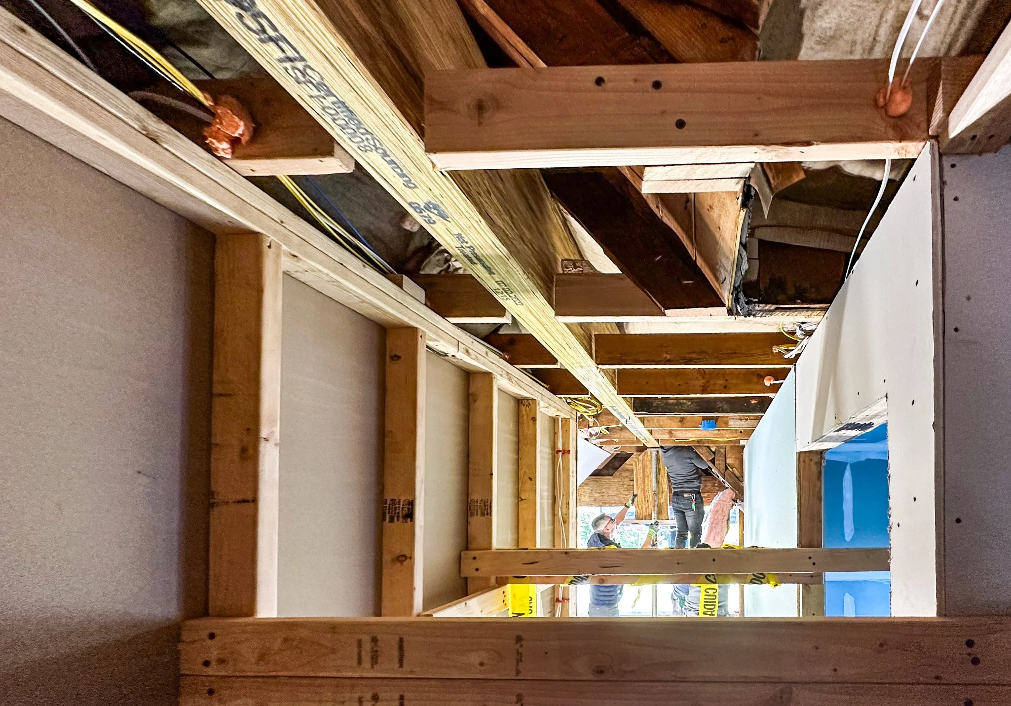 Interior view of a building under construction showing exposed wooden framing, insulation, drywall, and construction workers.