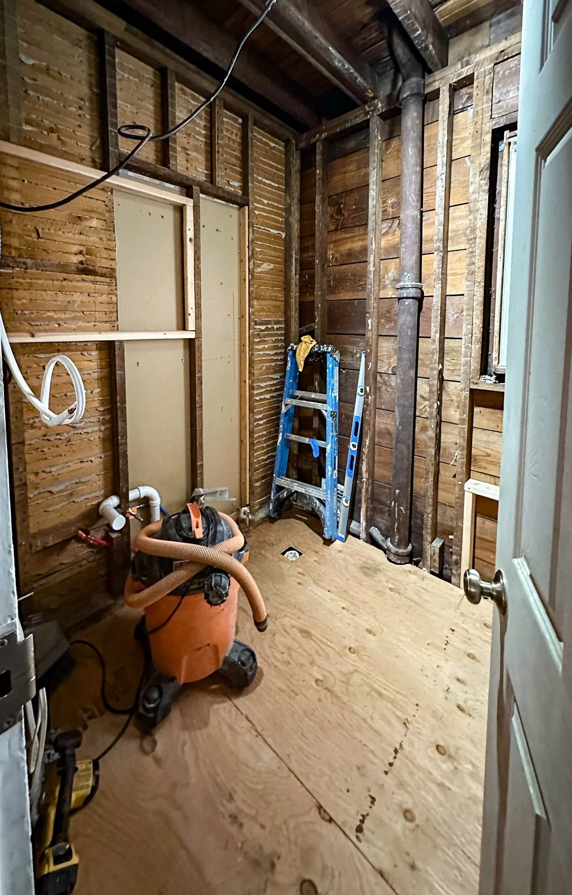 A small room under renovation with exposed wooden studs, a ladder, a wet/dry vacuum, and construction tools, with plywood flooring and unfinished walls.