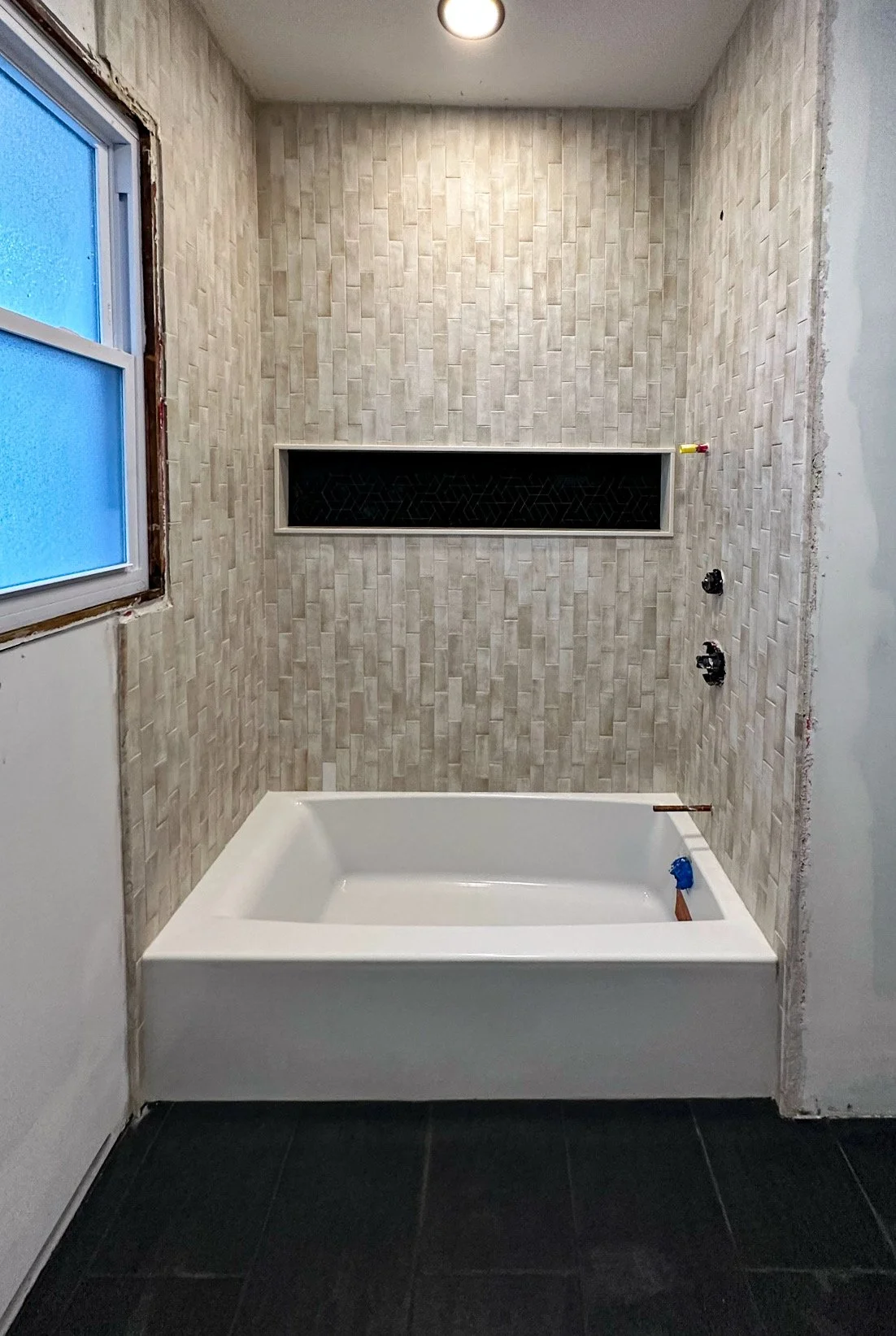 Bathroom under construction with a white bathtub, beige tiled wall, black recessed shelf, and a window with blue glass, no fixtures installed yet.