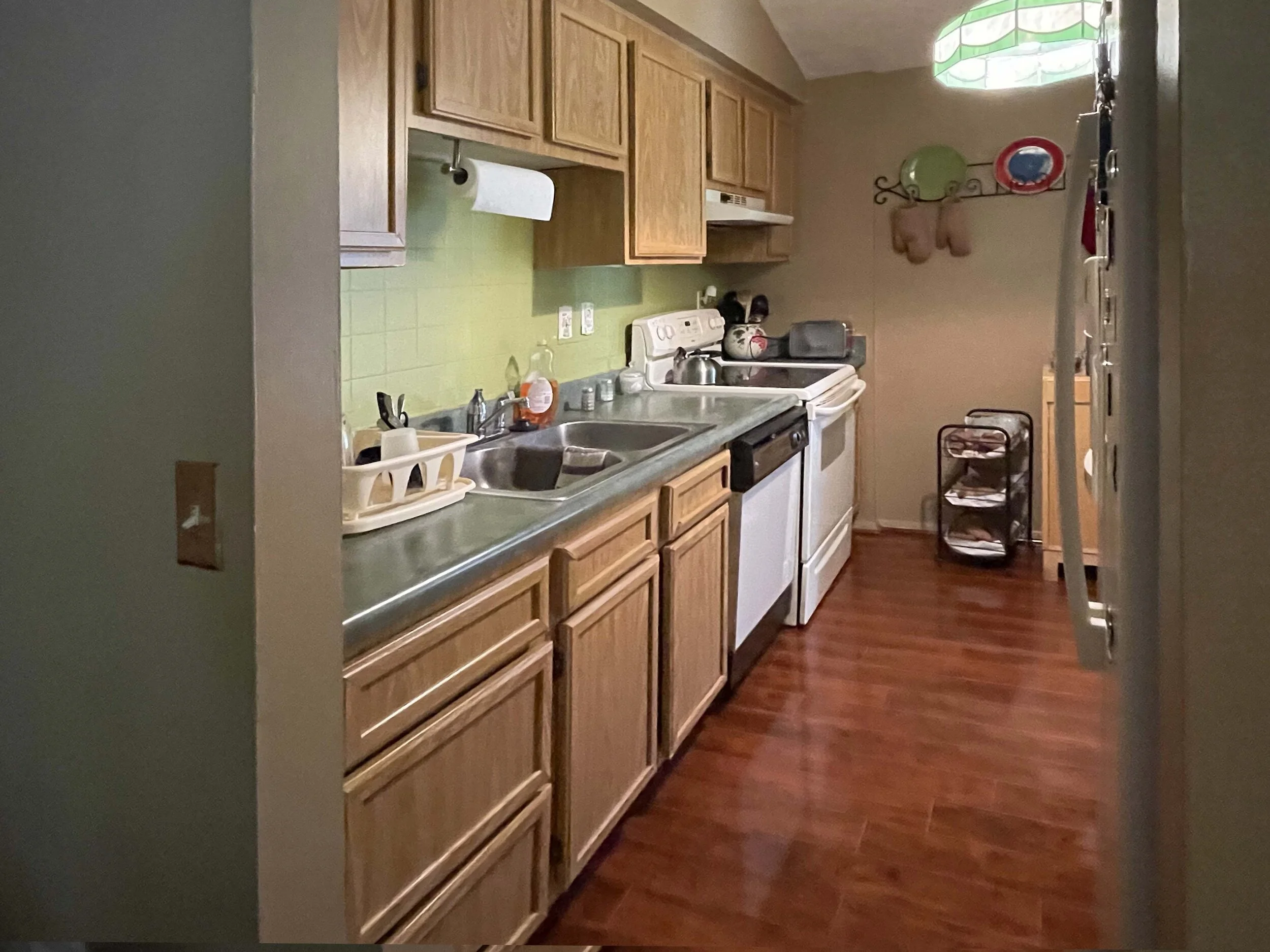 A kitchen with wooden cabinets, a green tile backsplash, and a white stove and sink. There are various kitchen items on the counter and a small black rolling cart with papers on it. The floor is wood, and a decorative light fixture hangs from the cei