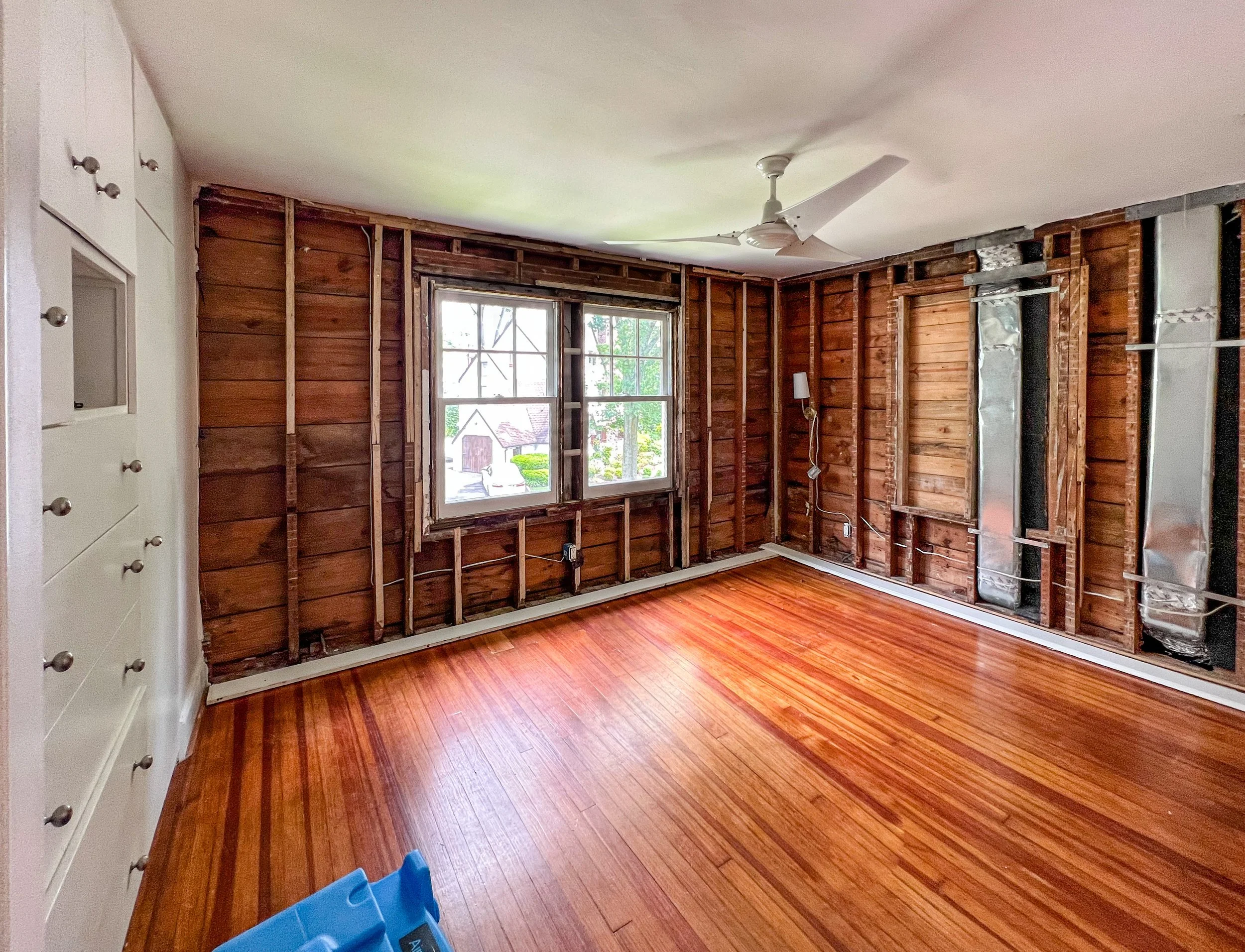 A room with all walls stripped to the studs, exposure to some ductwork and wiring, hardwood floors, a ceiling fan, a window showing an outdoor scene, and a partially painted built-in cabinet.