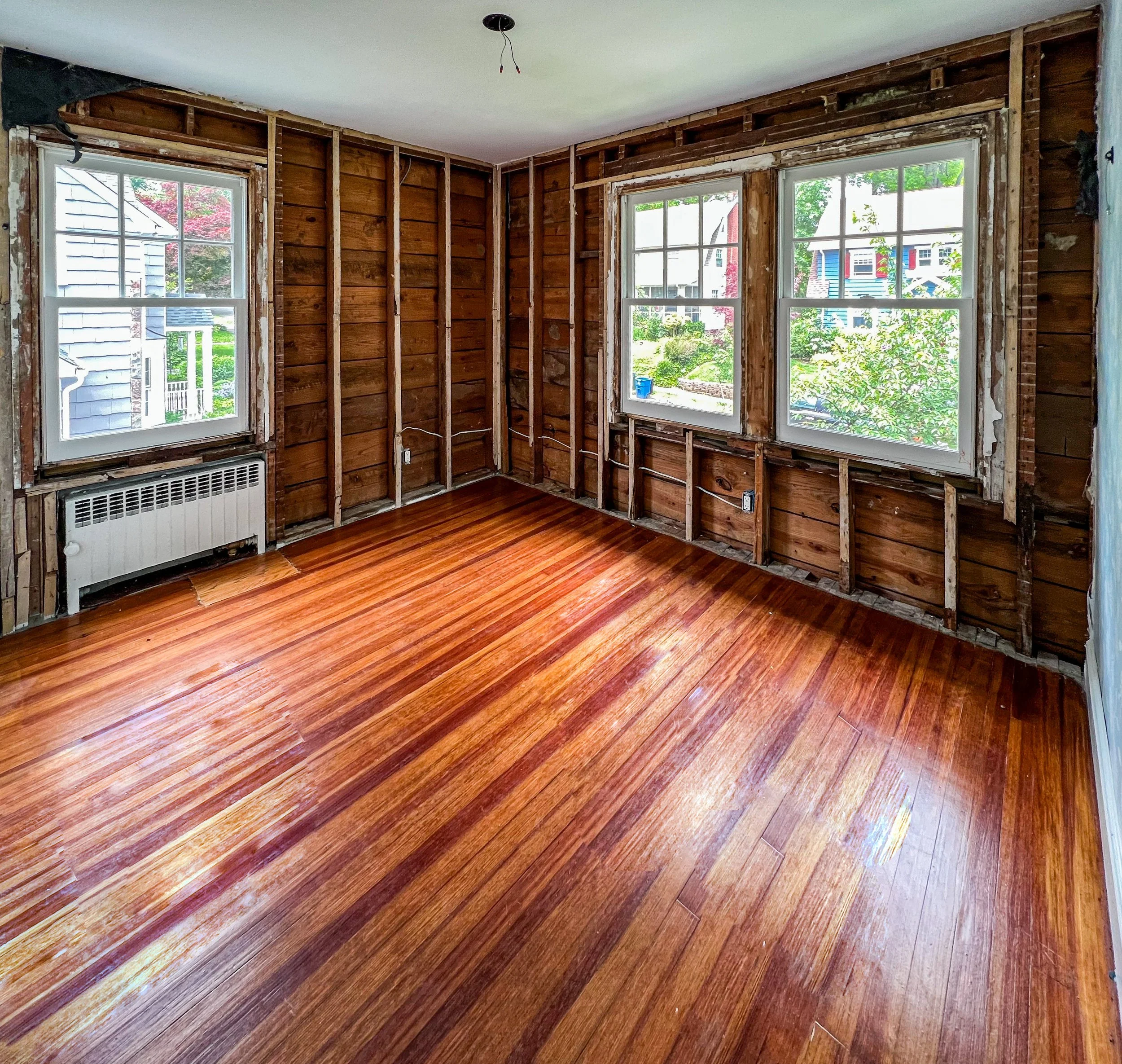 Empty room with wooden floors, three windows with white frames, and exposed wall studs and insulation, indicating ongoing renovation.