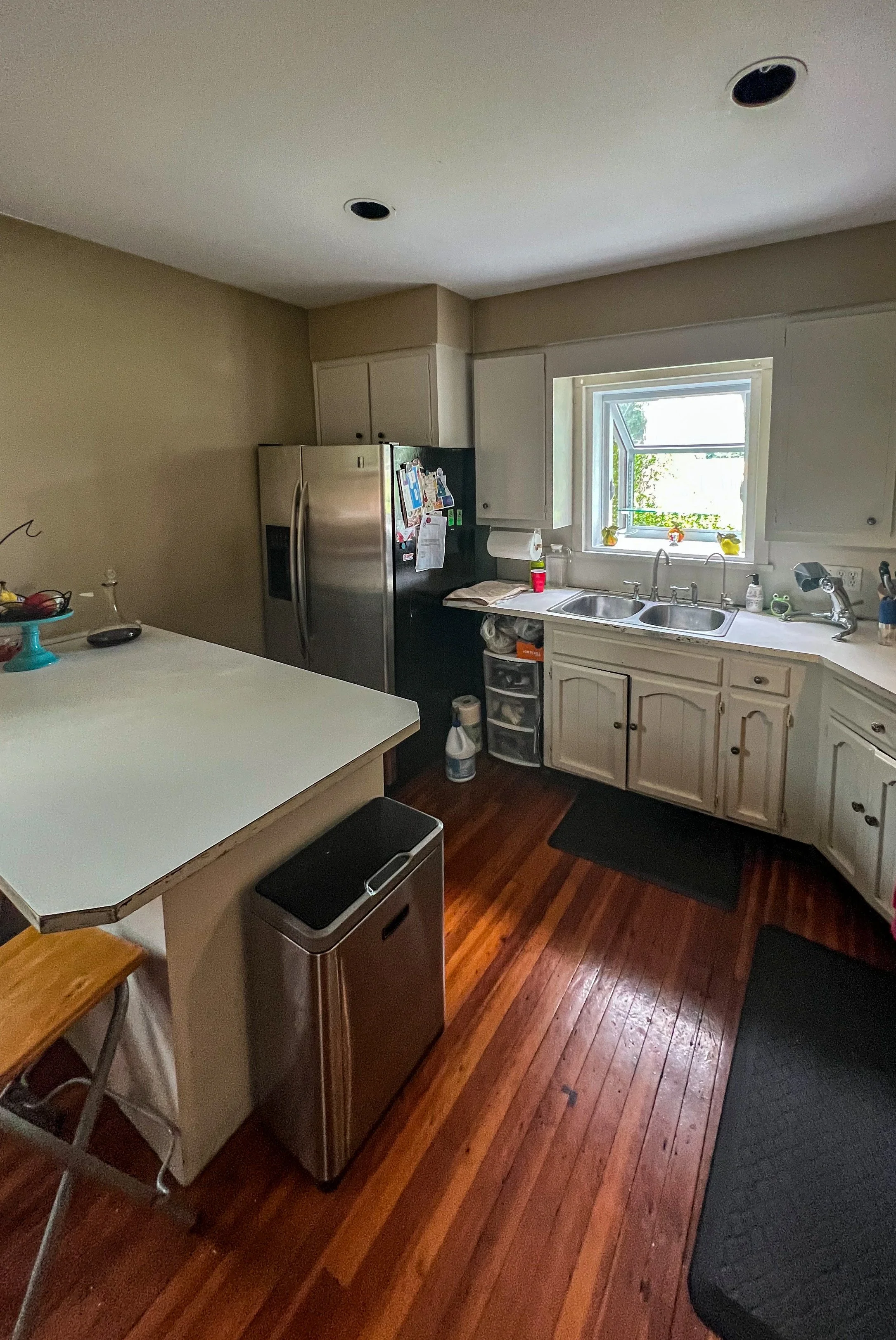 A kitchen with white cabinets, a window above the double sink, a stainless steel refrigerator with magnets, a trash can, hardwood floors, and black rugs.