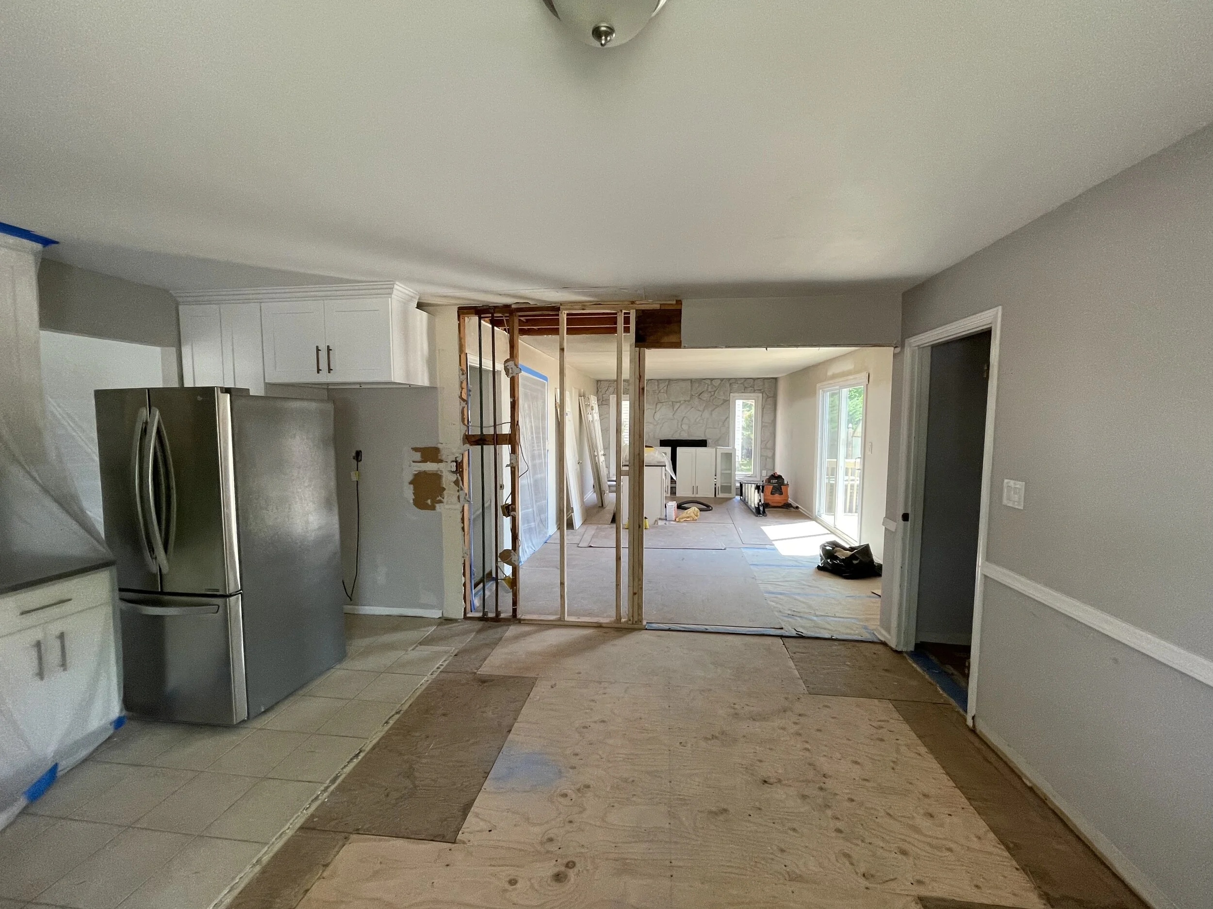 Interior of a house under renovation, showing a kitchen with a stainless steel refrigerator, white cabinets, and an open wall framing leading to a living room area with a stone fireplace and large windows.