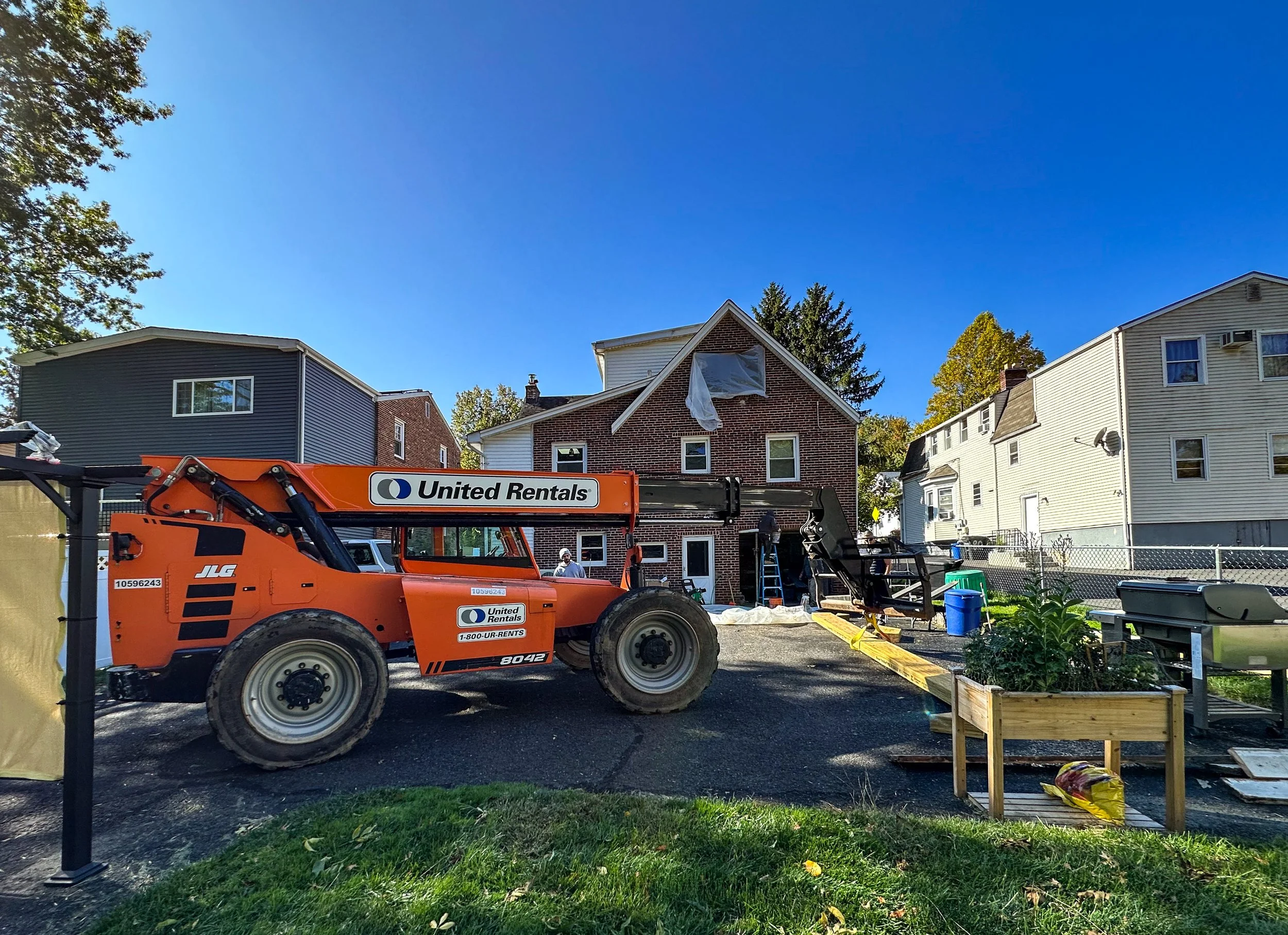 Construction crew working on a house renovation with a large orange United Rentals crane and construction materials on site.
