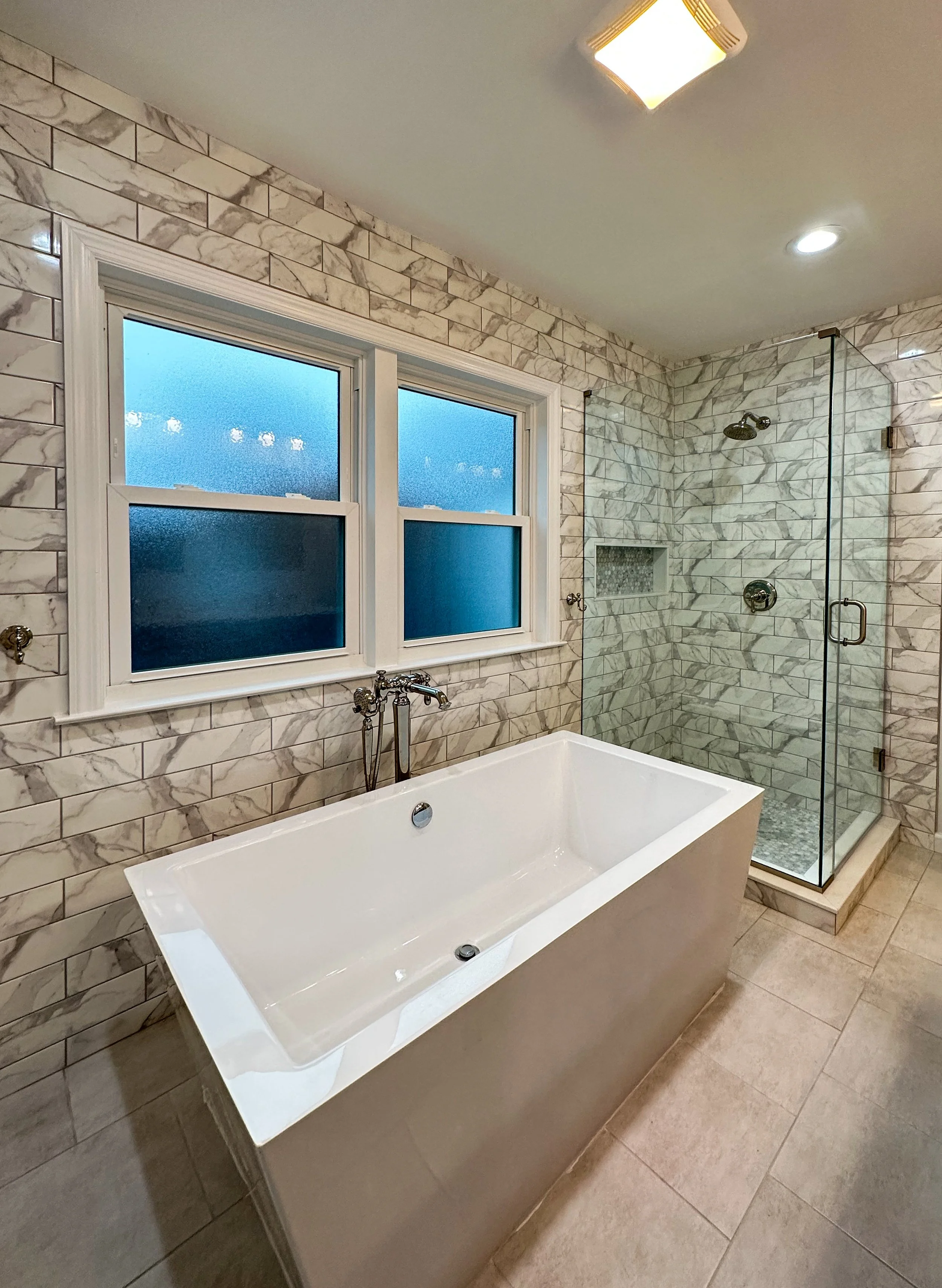 Bathroom with white bathtub, glass-enclosed shower, and tiled walls decorated with gray and white marble patterns.