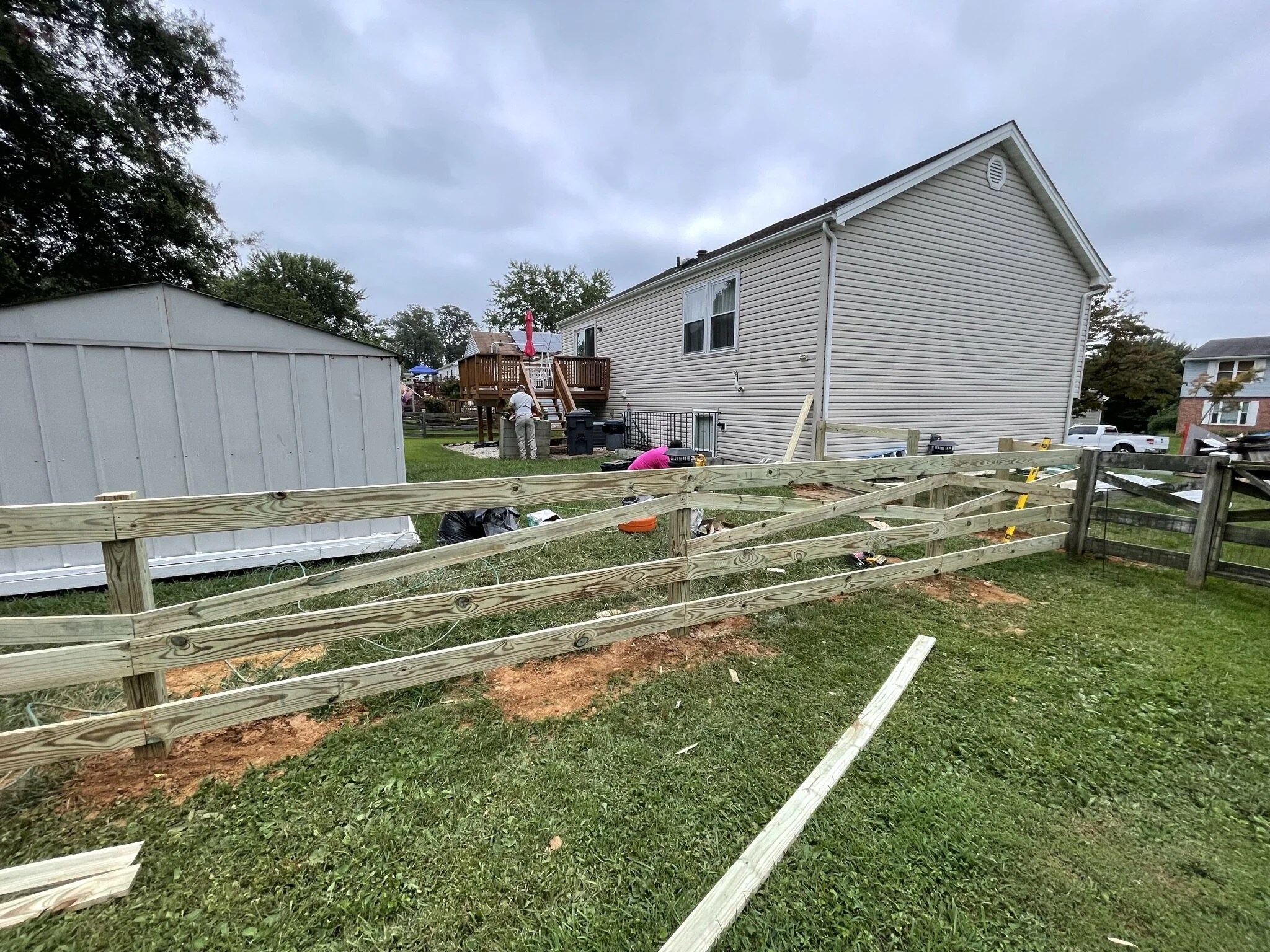 Backyard with partially built wooden fence, a shed, and a house with a deck. Several people are working, and construction tools are visible.