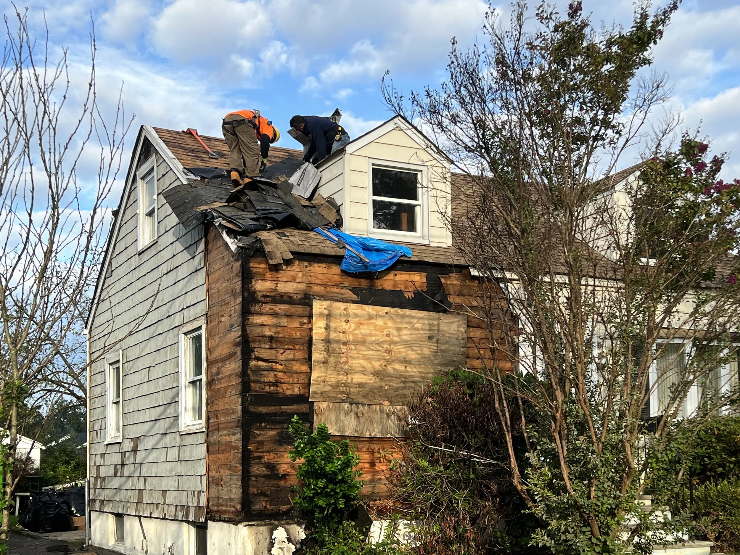 Two workers repairing a damaged roof on a multi-story house, with some shingles removed, during daytime with partly cloudy sky.